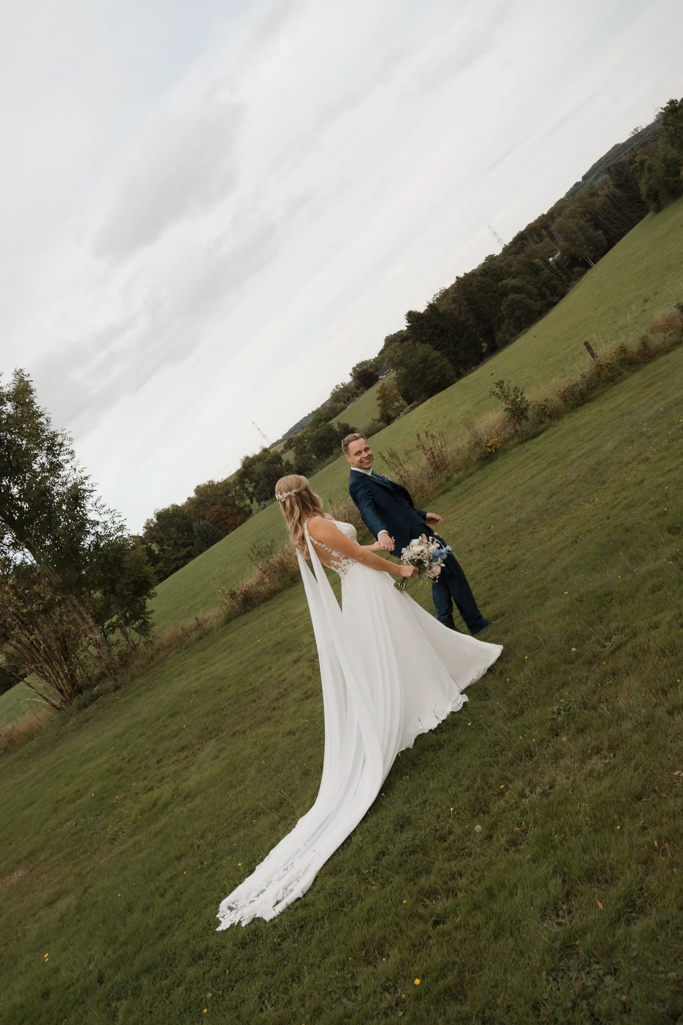 Ein Brautpaar hält Händchen im Grünen, die Braut trägt ein weißes Hochzeitskleid und die(zumindest) auf dem Foto sind glückliche Gesichter zu sehen, im Hintergrund sind Bäume und ein bewölkter Himmel.