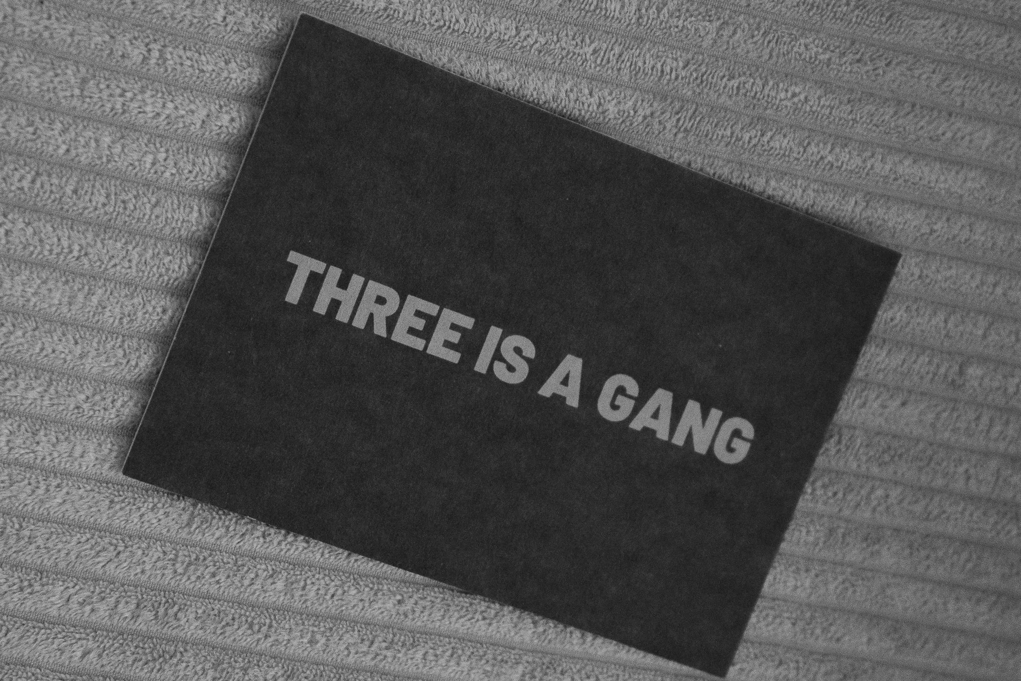 Ein schwarzes Plakat mit weißen Buchstaben, auf dem steht 'THREE IS A GANG', liegt auf einer rautenförmigen, gestreiften Oberfläche.