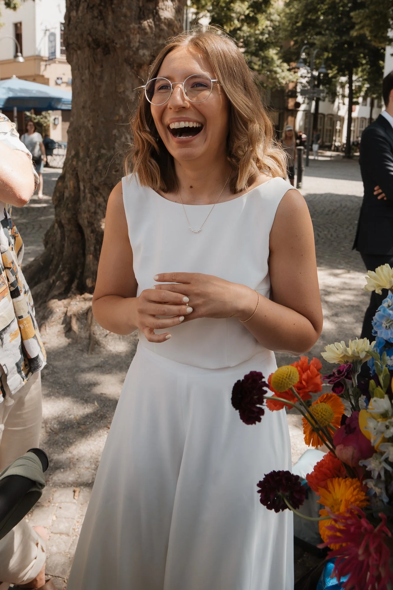 Eine lachende Frau in einem weißen Kleid, wahrscheinlich bei einer Feier im Freien, mit bunten Blumen im Vordergrund und einem Baum im Hintergrund.