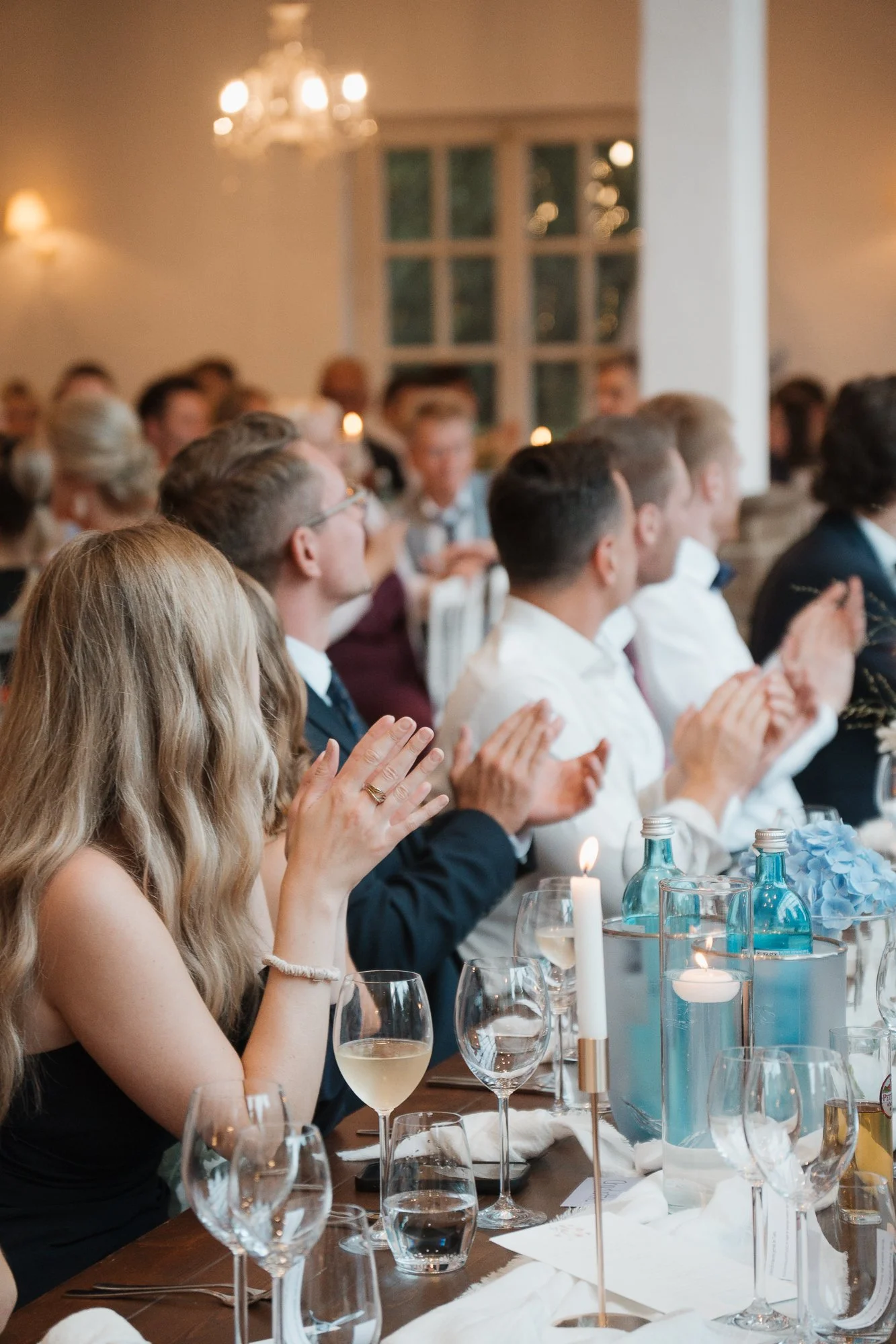 Eine Gruppe von Menschen sitzt bei einer formellen Veranstaltung, die Tische sind mit Kerzen, Blumen und Gläsern gedeckt, die Gäste applaudieren.