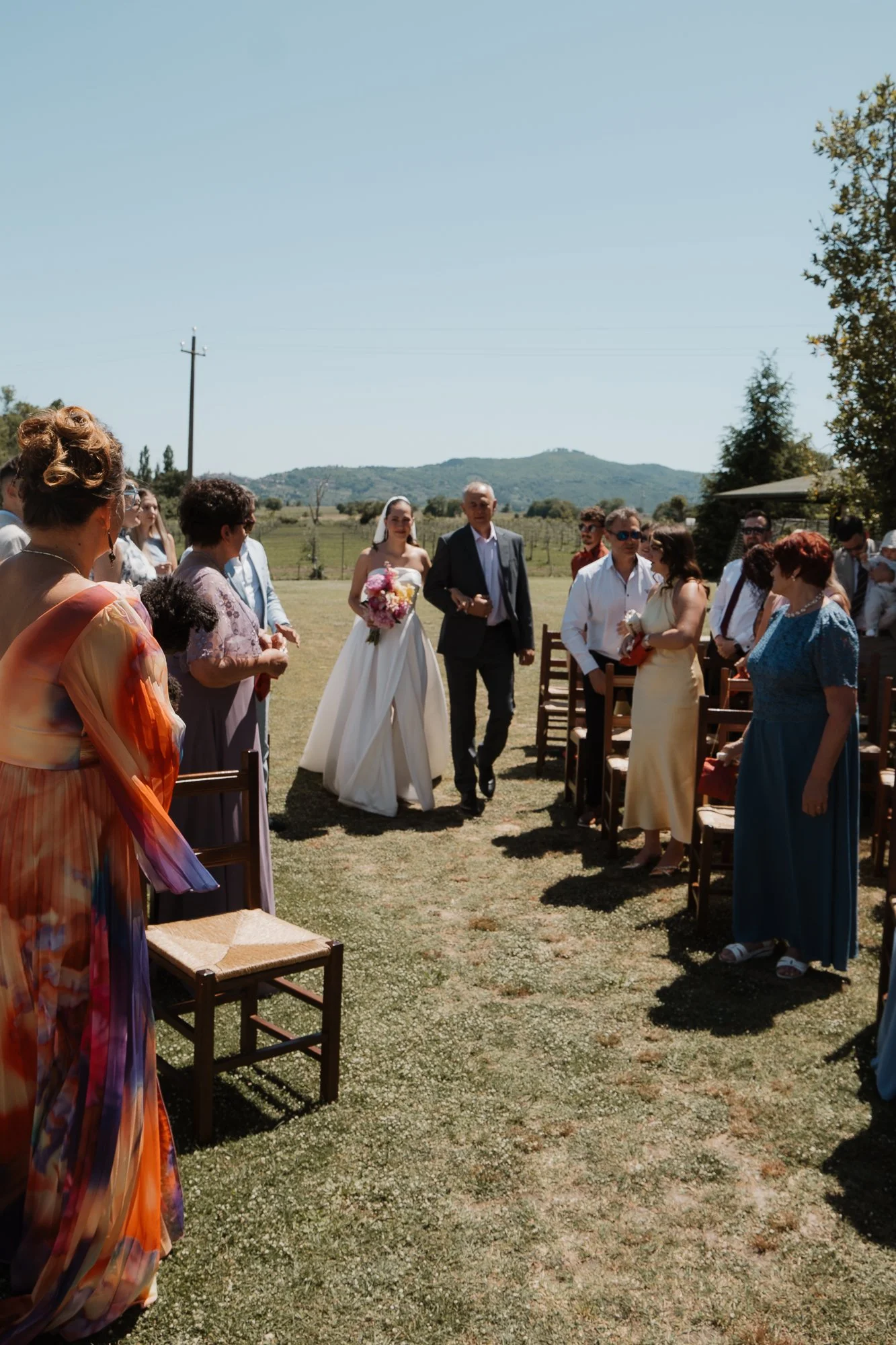 Eine Hochzeitszeremonie im Freien mit einer Braut im weißen Kleid, die von einem Mann begleitet wird, umgeben von Gästen, auf einer Wiese bei Sonnenschein, mit Bäumen und Hügel im Hintergrund.