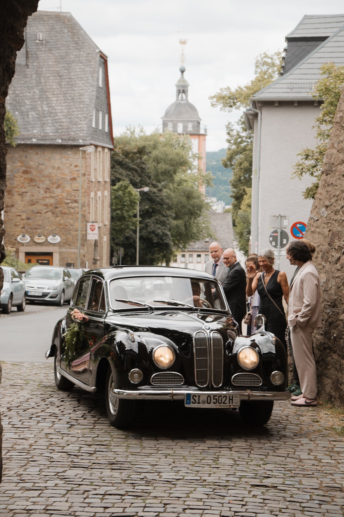 Eine schwarze klassische Limousine steht auf einer gepflasterten Straße, umgeben von Menschen in formeller Kleidung, wahrscheinlich bei einer Hochzeitsfeier. Im Hintergrund sind alte Gebäude und ein Kirchturm sichtbar.