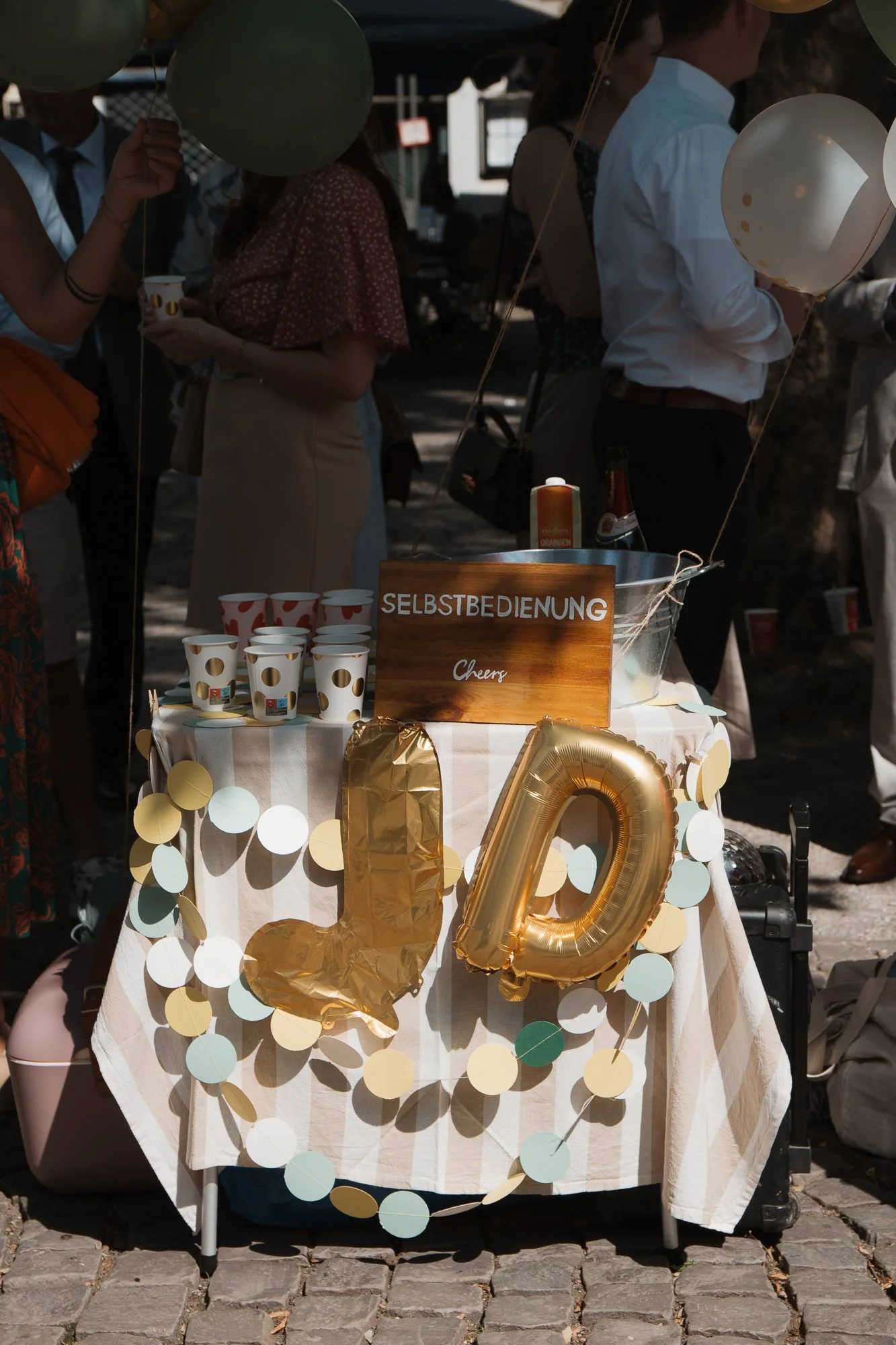 Tisch mit goldenen Ballons, Pappbecher, und einem Schild mit der Aufschrift 'SELBSTBEDIENUNG'.