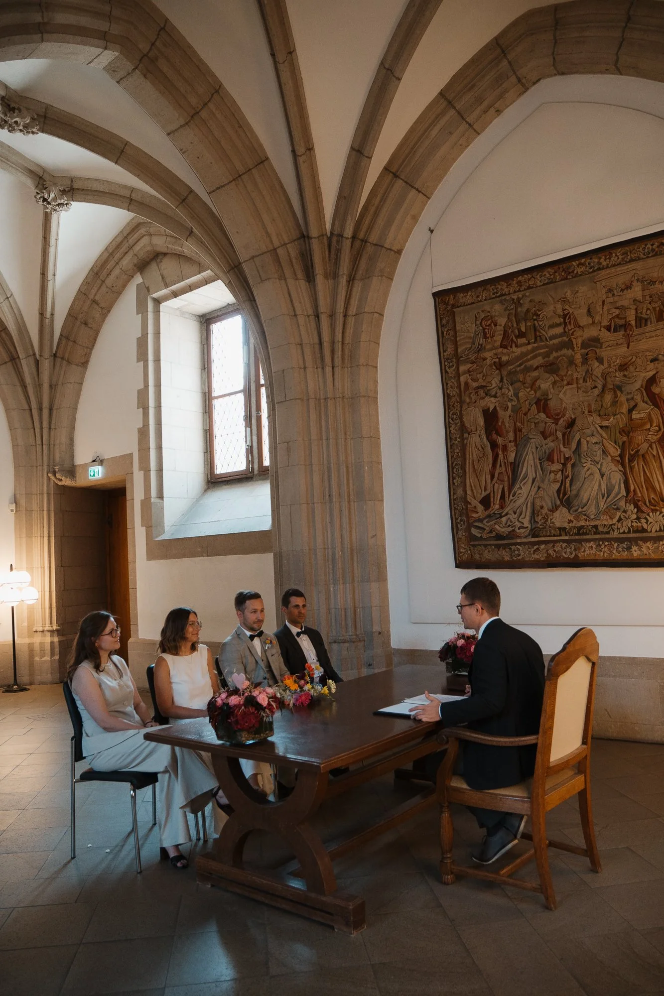 Eine Trauung in einem gotischen Raum mit hohen Bögen, großen Fenstern und einem Wandteppich zeigt fünf Menschen, vier von ihnen sitzen an einem langen Tisch mit Blumen, während der fünfte Mensch, der Bräutigam, vor ihnen sitzend die Zeremonie durchfü