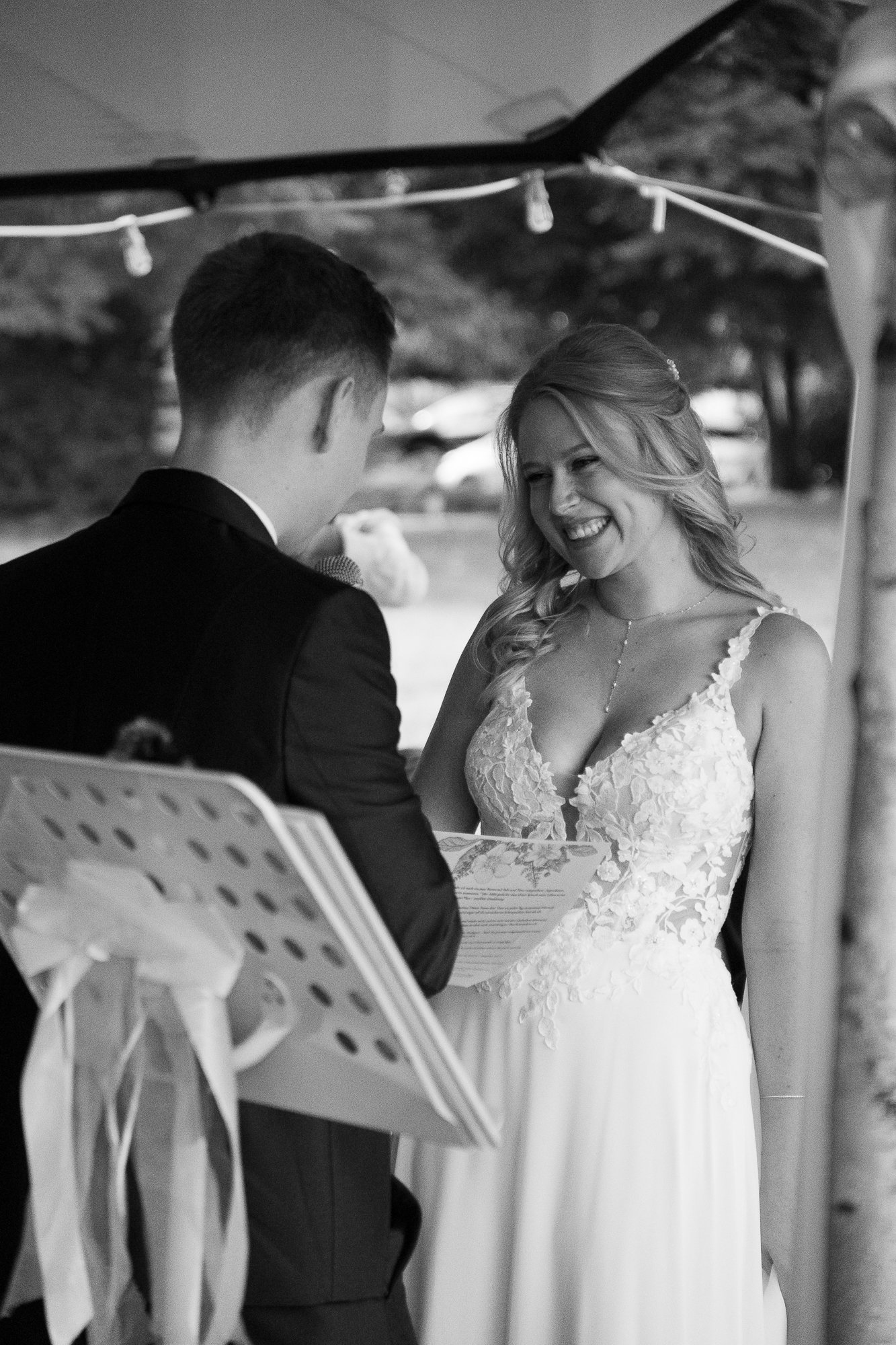 Braut und Bräutigam bei einer Zeremonie im Freien, die Braut trägt ein weißes Hochzeitskleid mit Blumenmuster, der Bräutigam trägt einen schwarzen Anzug, beide lachen.