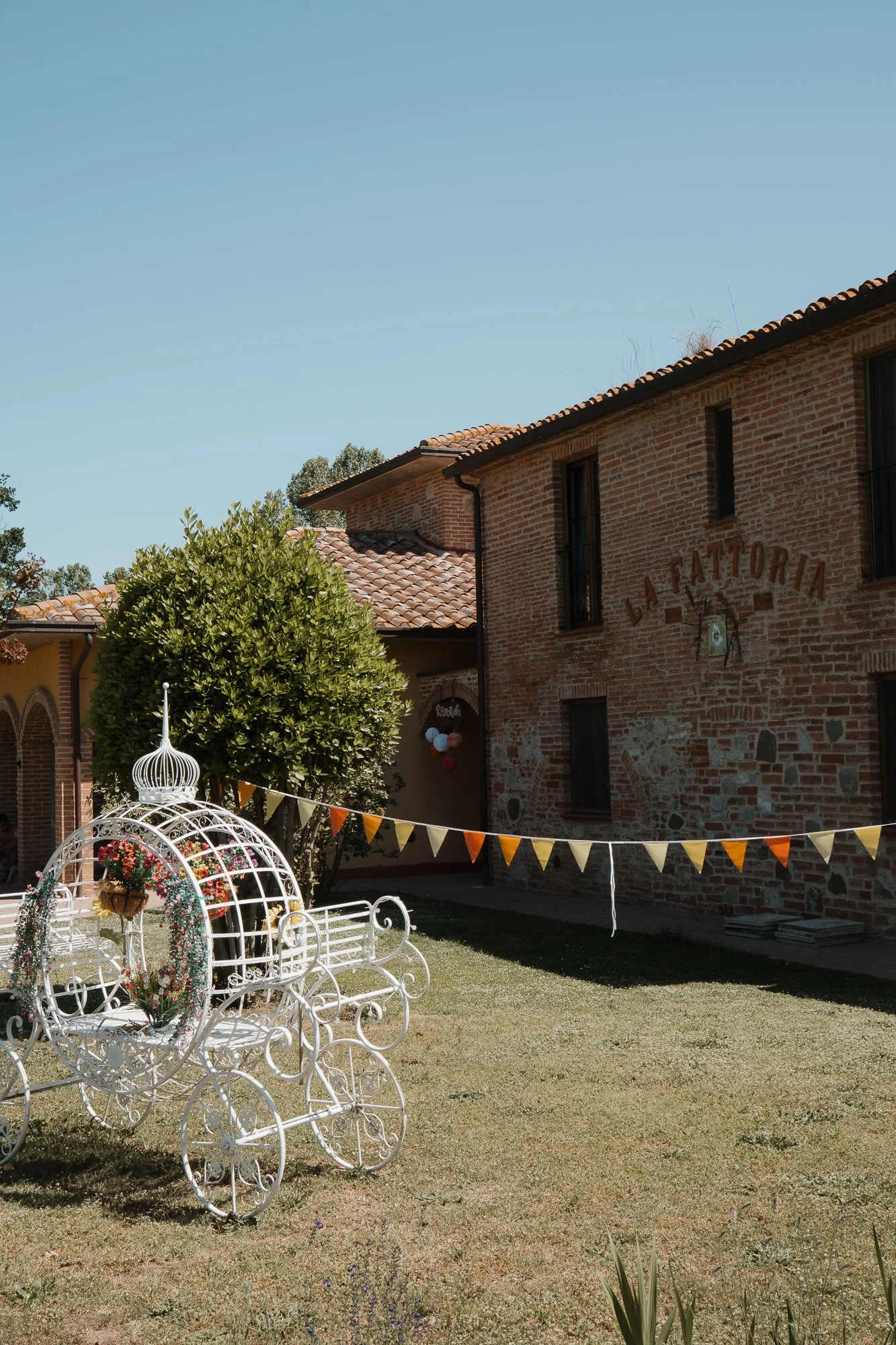 Ein outdoor Fest mit einem weißen, dekorativen Kutschen-Träger in einem Garten, bunten Fähnchen und einer großen Büsche, im Hintergrund ein rotes Backsteingebäude mit dem Schriftzug "LA FATTORIA".