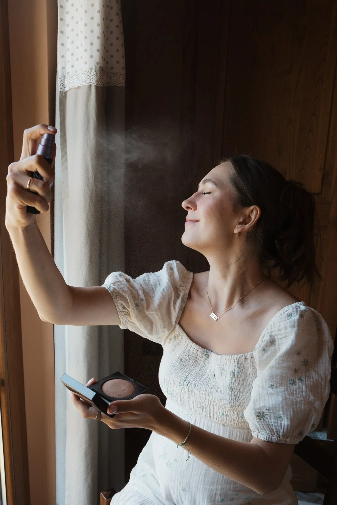 Frau sprüht Parfüm auf ihr Gesicht, während sie mit einem Kompaktpuder in der Hand posiert.