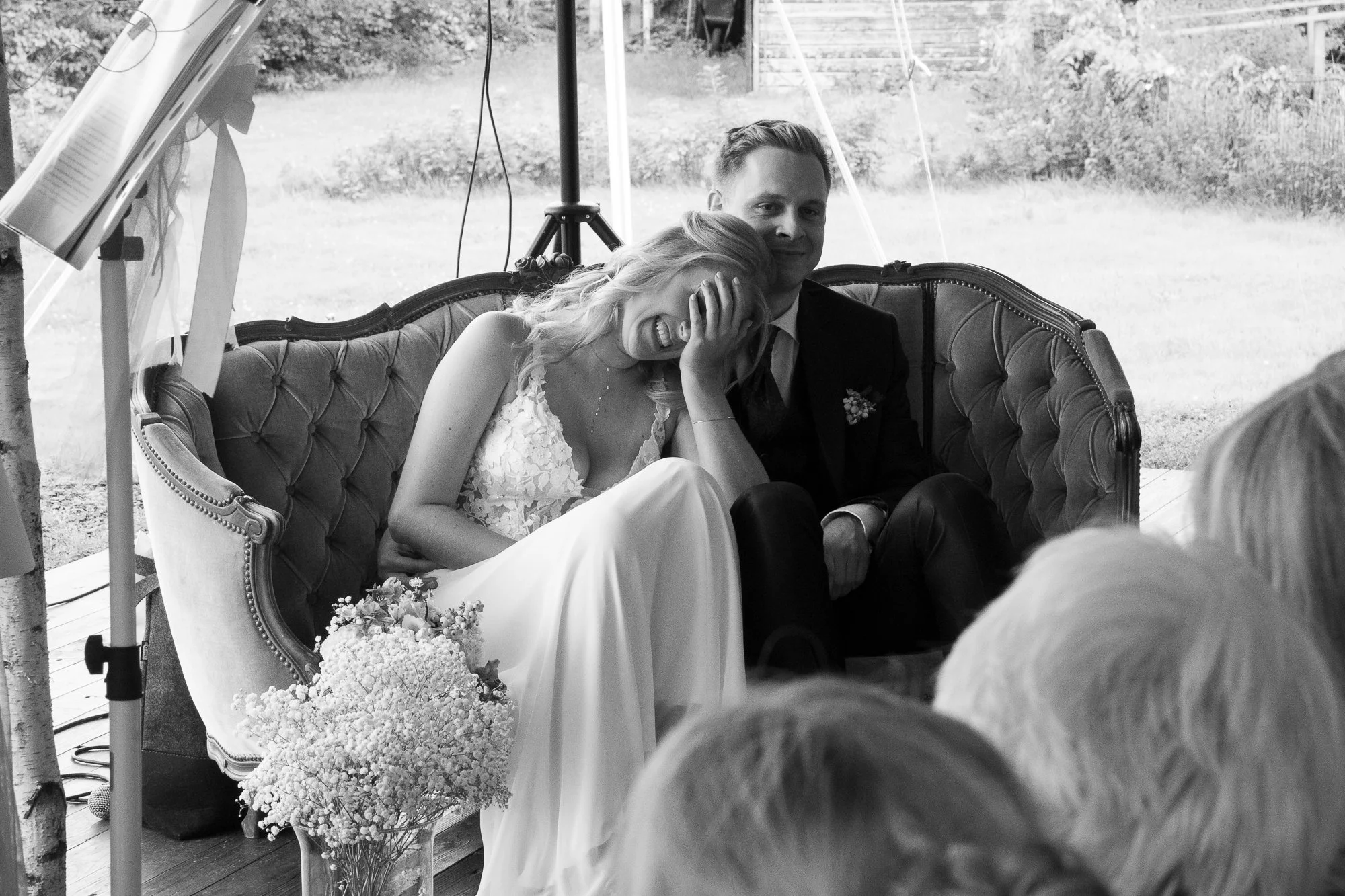 Ein Brautpaar sitzt lachend auf einer alten Couch während einer Hochzeit. Die Braut trägt ein weißes Kleid mit Spitze, der Bräutigam einen dunklen Anzug. Sie scheinen inmitten einer Unterhaltung mit Gästen zu sein, mit einer natürlichen, grünen Lands