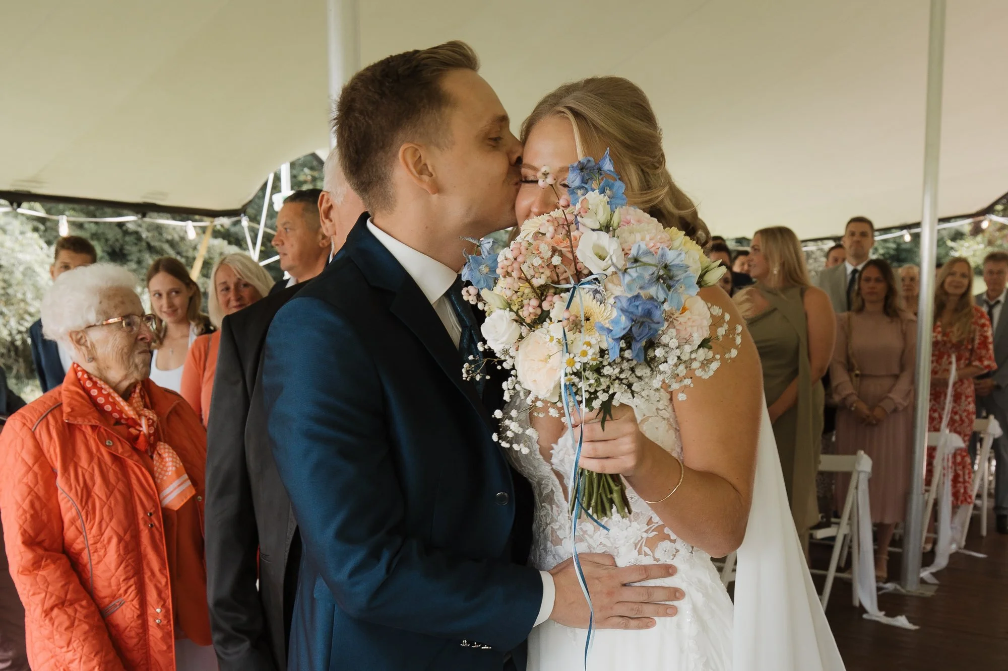 Ein Brautpaar bei ihrer Hochzeit, das sich küsst, umgeben von Gästen in einem Festzelt.