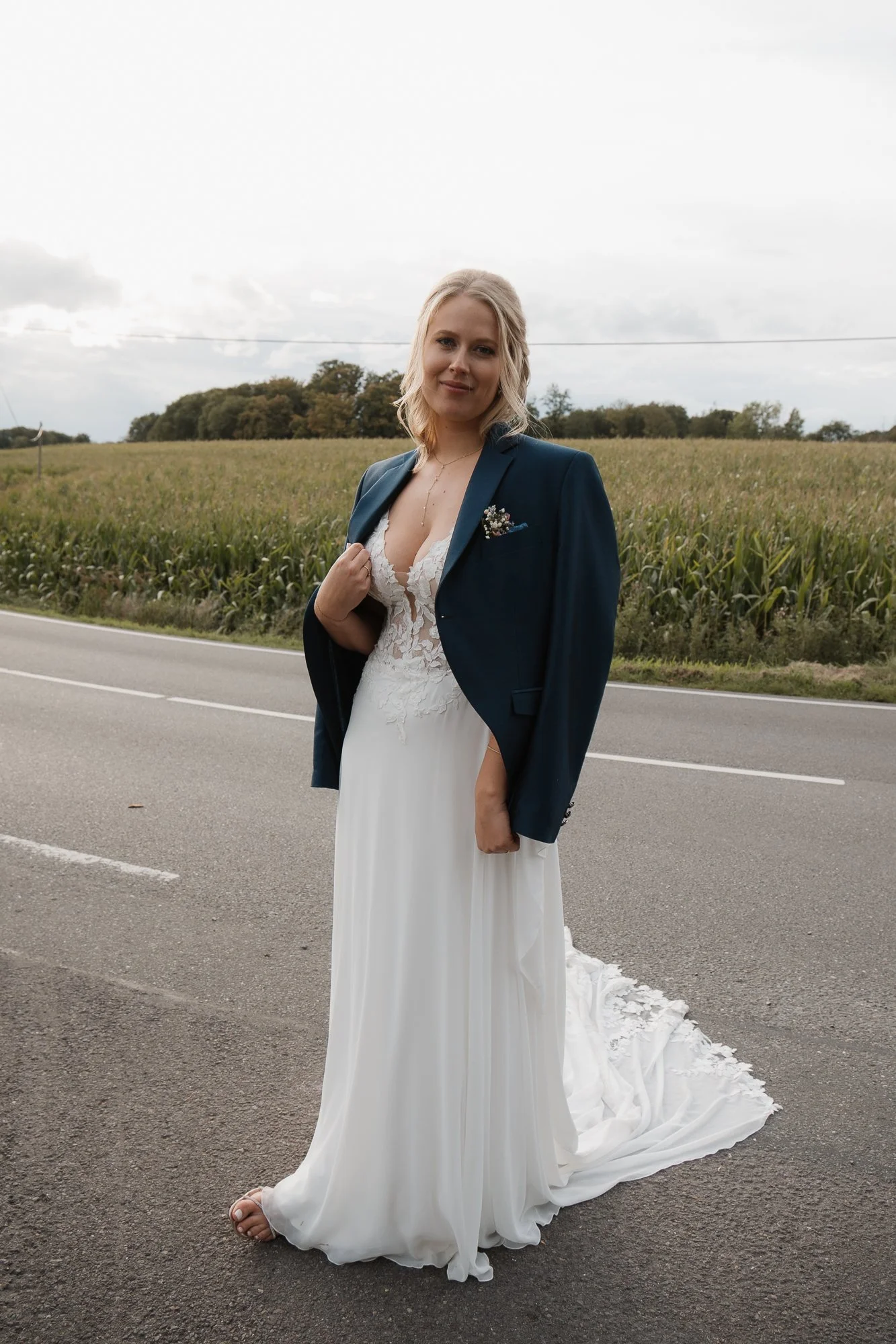 Braut steht auf einer Straße, trägt ein weißes Hochzeitskleid und eine dunkle Jacke, im Hintergrund Felder und Wolkenhimmel.