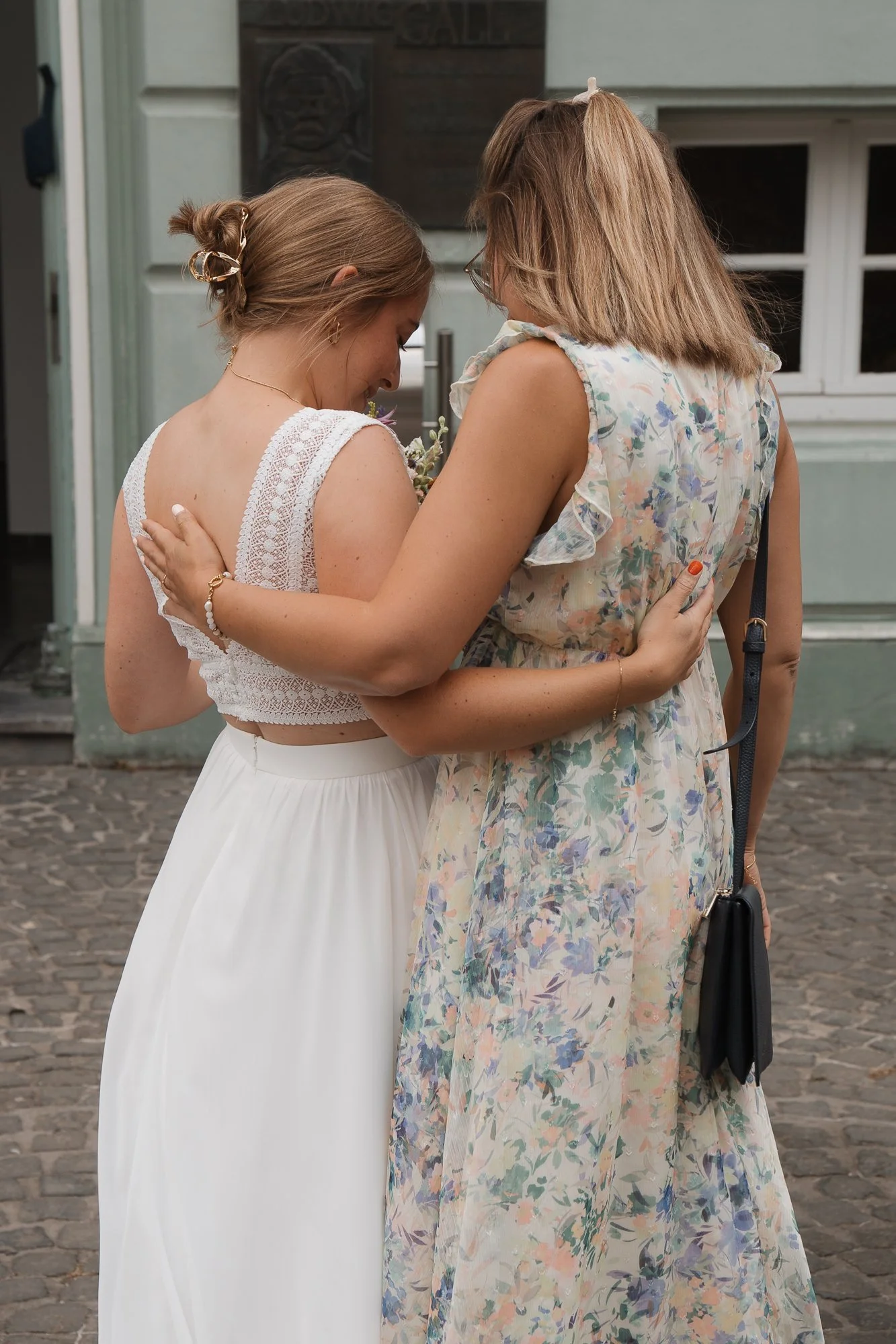 Zwei Frauen umarmen sich herzlich auf einer Kopfsteinpflasterstraße, eine trägt ein weißes Brautkleid, die andere ein bunt bedrucktes Kleid.