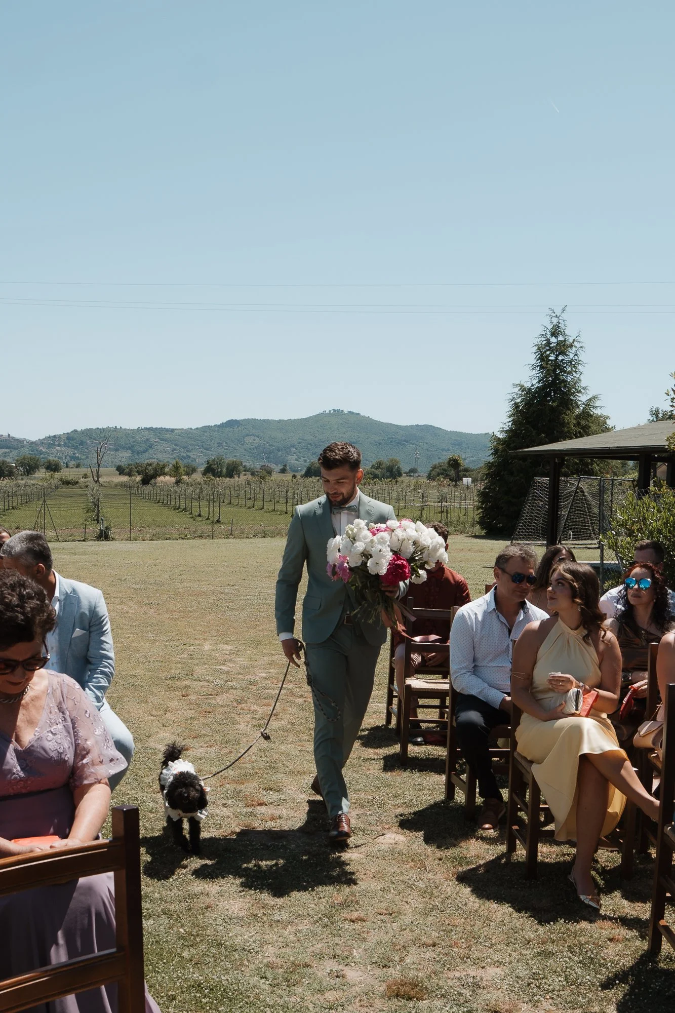 Ein Hochzeitsempfang im Freien mit einem Mann, der einen Blumenstrauß trägt, und einer Gruppe von Gästen, die auf Stühlen sitzen. Einige Gäste tragen Sonnenbrillen, im Hintergrund sind Weinberge und Berge sichtbar.