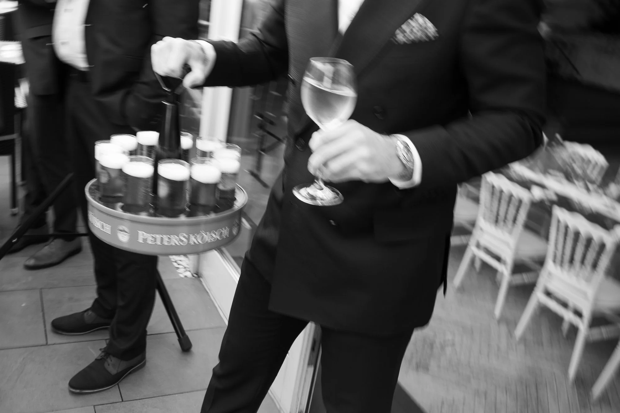 Ein Mann in formeller Kleidung hält ein Glas Wein in einer Hand und steht neben einem Tablett mit Flaschen und Gläsern. Mehrere Stühle sind im Hintergrund sichtbar.