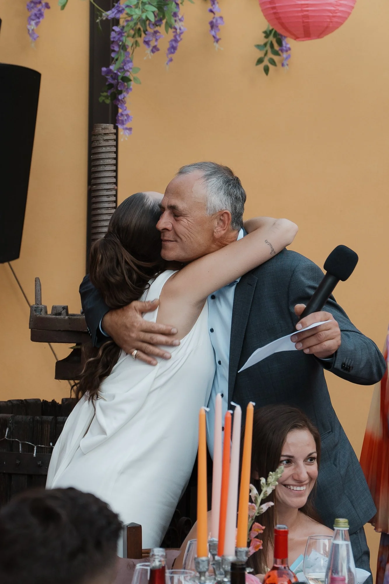 Ein Mann umarmt eine Frau bei einer Feier, während er ein Mikrofon hält, im Vordergrund sitzt eine lachende Frau an einem Tisch mit Kerzen.