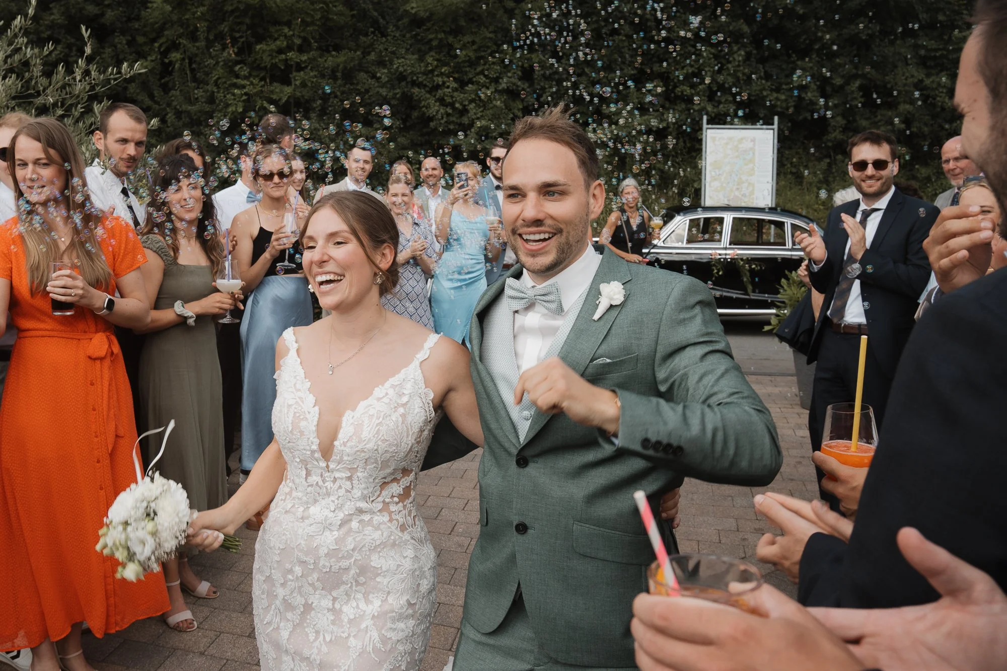 Ein frisch verheiratetes Paar, die Braut im weißen Spitzenkleid, der Bräutigam in einem grauen Anzug, bei ihrer Hochzeit. Sie werden von Freunden und Familie umgeben, die im Hintergrund bei sonnigem Wetter feiern und Blasen pusten.