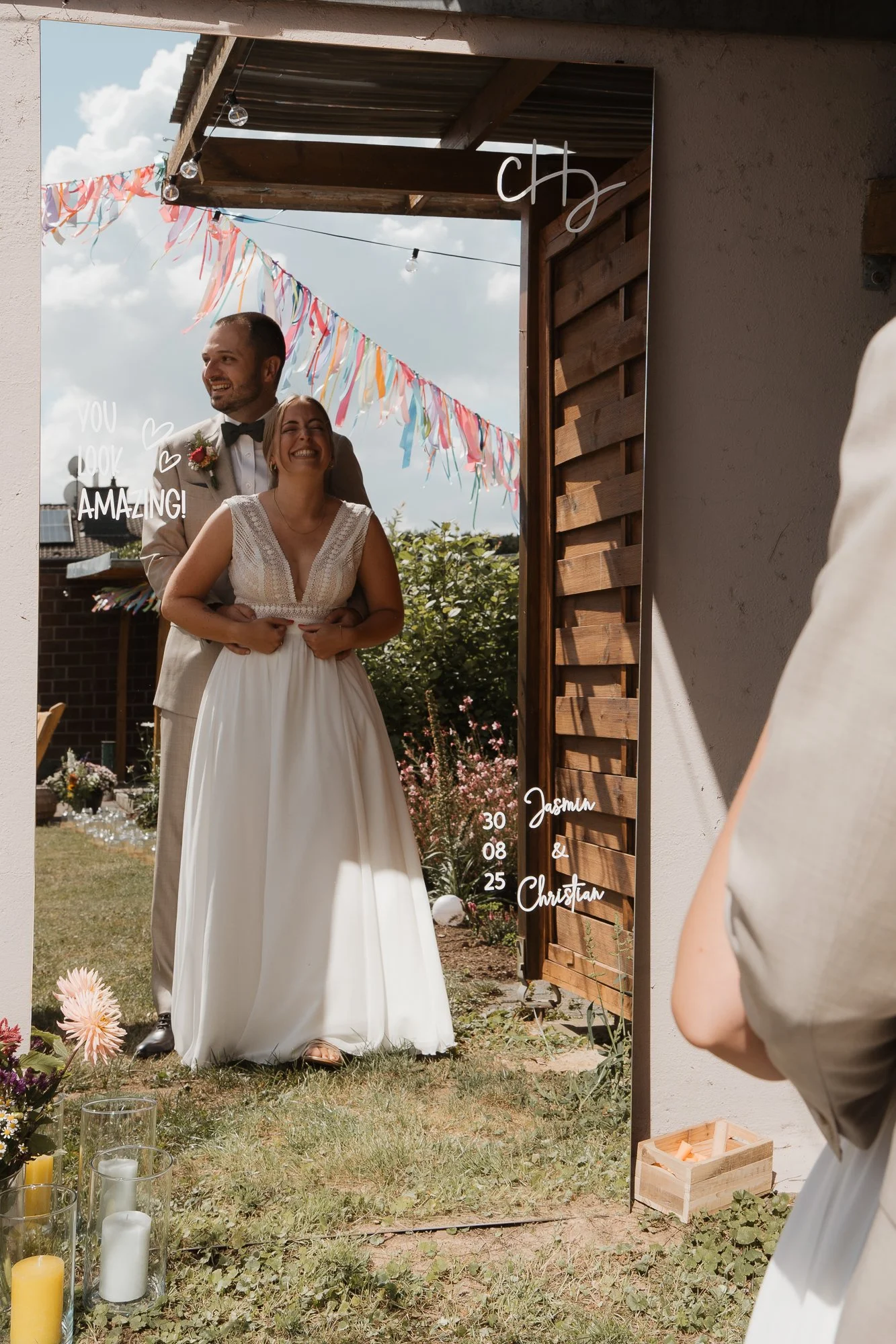 Ein Mann und eine Frau in Hochzeitskleidung stehen vor einem Spiegel im Garten, während eine Person im weißen Kleid das Foto macht. Es gibt bunte Girlanden im Hintergrund, Blumen und Kerzen auf dem Boden.