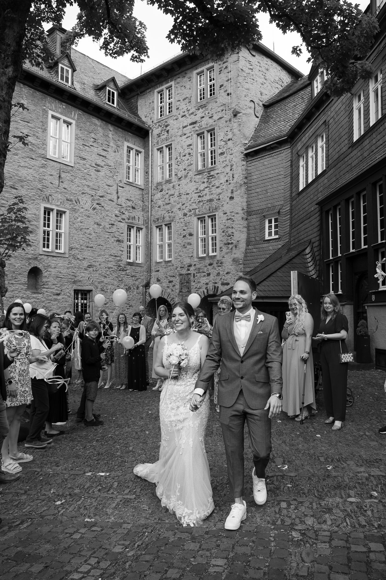 Ein Brautpaar hält Händchen und geht bei einer Hochzeitsfeier. Die Braut trägt ein langes, verziertes Hochzeitskleid und hält einen Blumenstrauß. Der Bräutigam trägt einen Anzug mit Fliege und weiße Turnschuhe. Freunde und Familie im Hintergrund feie