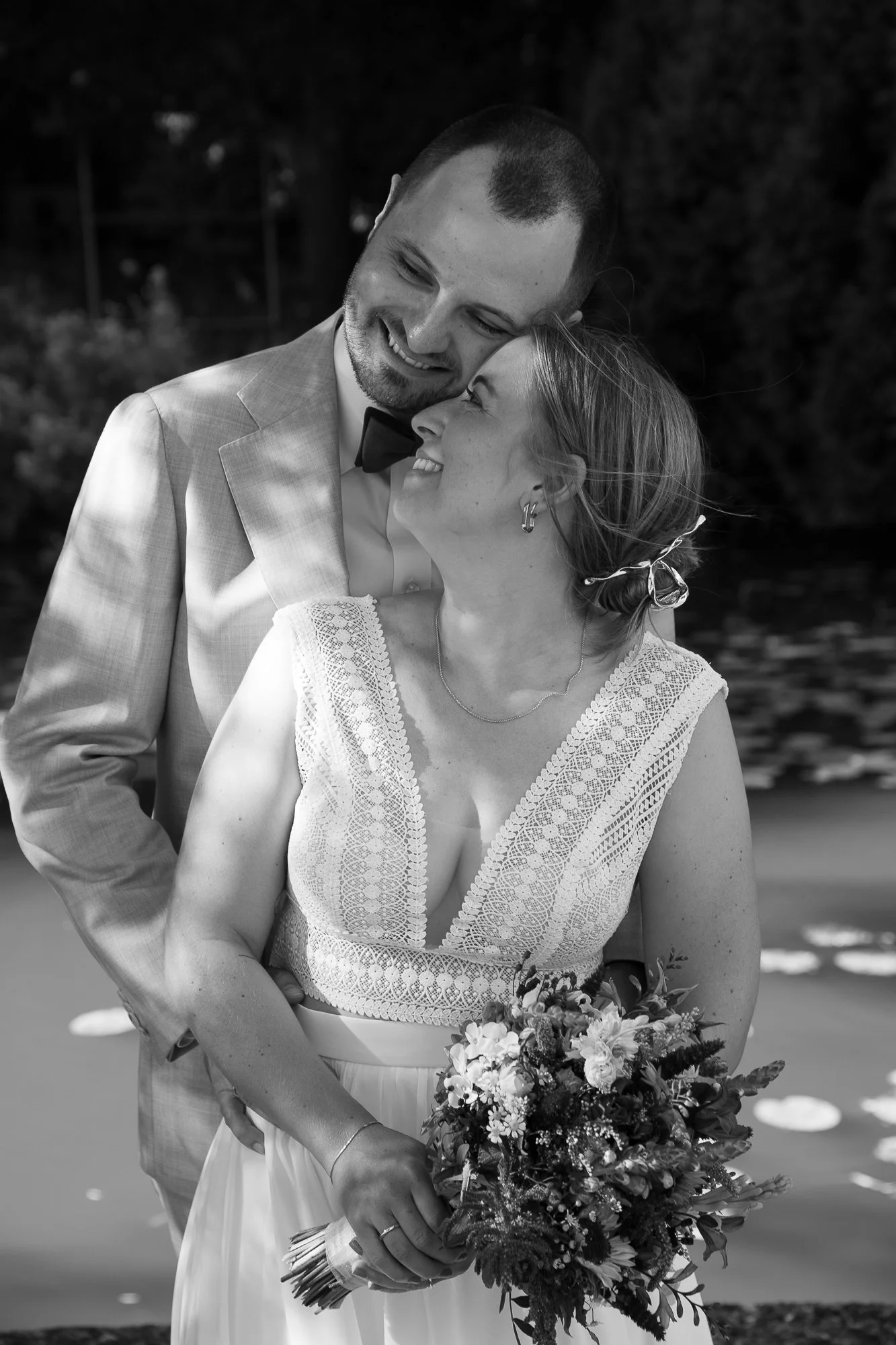 Ein glückliches Brautpaar bei ihrer Hochzeit, der Mann trägt einen Anzug mit Fliege, die Frau hält einen Blumenstrauß, sie schauen sich liebevoll an, im Hintergrund Wasser und Bäume.