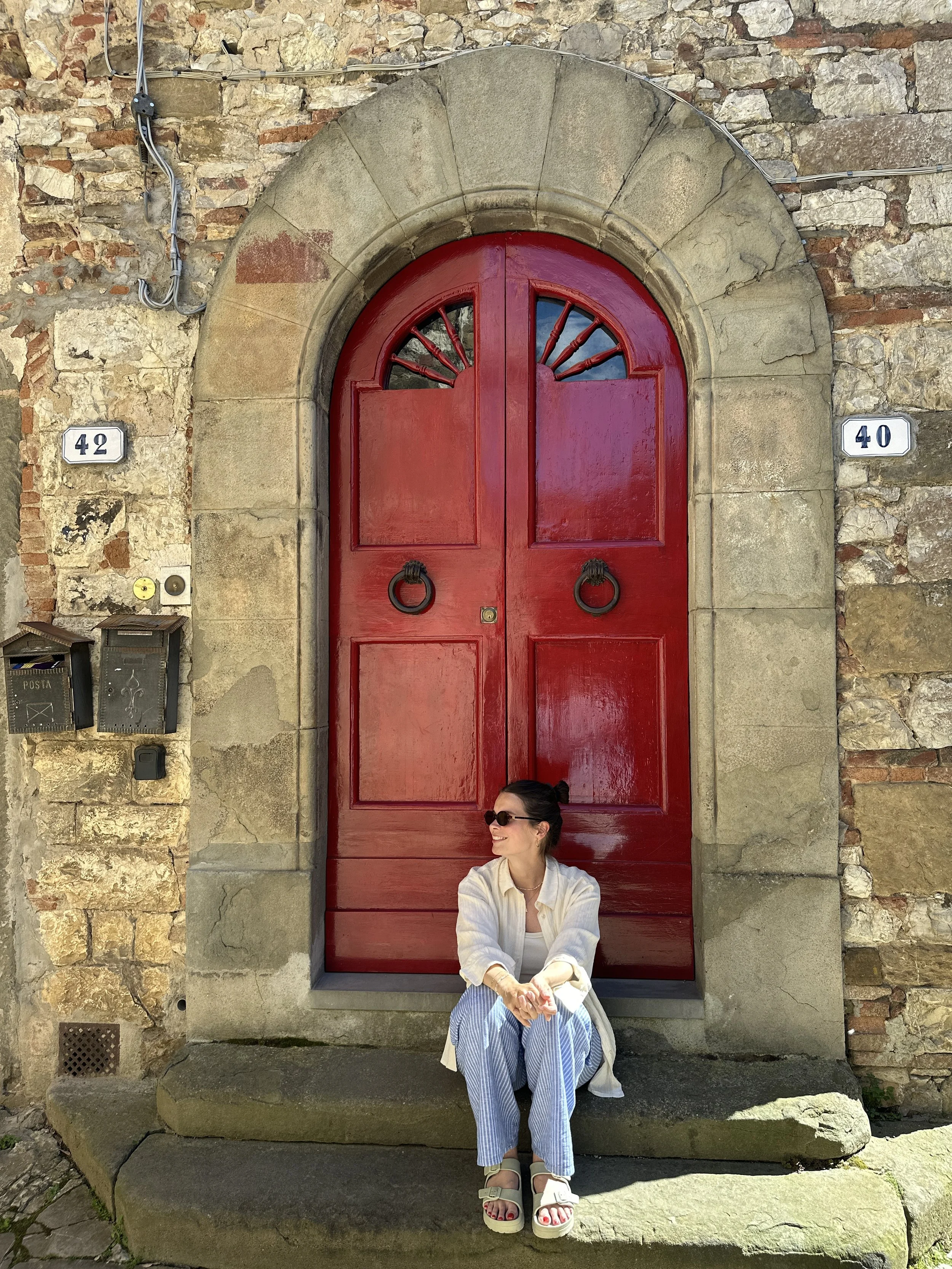 Eine Frau sitzt auf einem Stein vor einer roten Holztür in einer Steinmauer, trägt Sonnenbrille, weiße Jacke, blaue gestreifte Hosen und Sandalen.