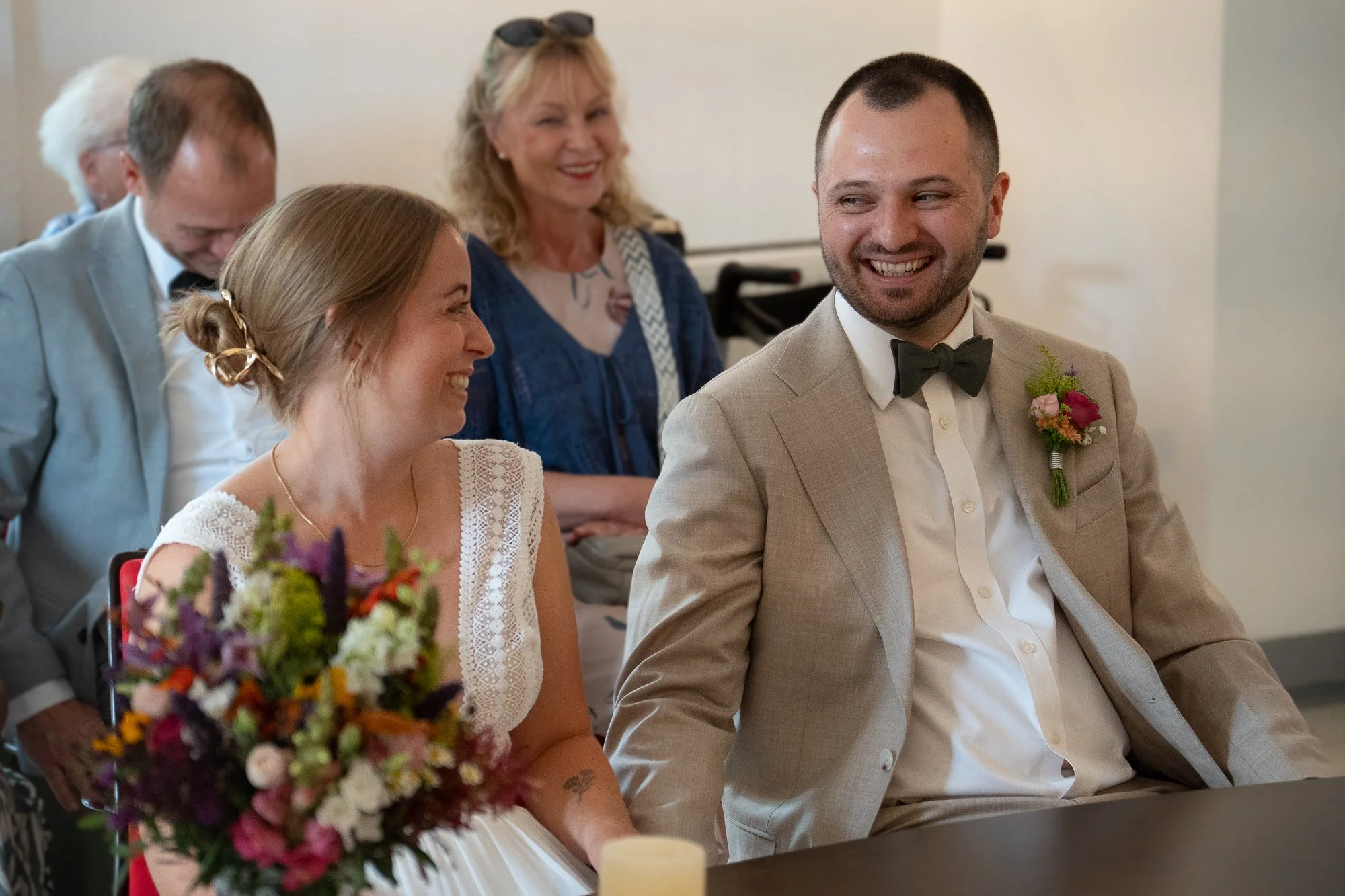 Ein glückliches Brautpaar bei einer Hochzeit, die Braut trägt ein weißes Kleid und hält einen bunten Blumenstrauß, der Bräutigam trägt einen hellbeigen Anzug mit schwarzen Fliege und ein rotes und grünes Blumenboutonniere. Im Hintergrund lachen und f