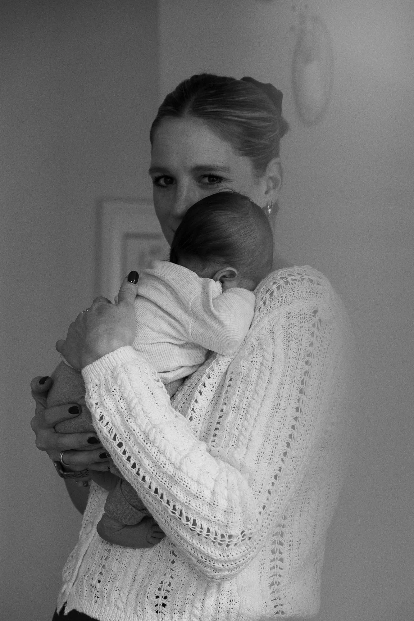 Eine Frau hält ein schlafendes Baby im Arm, beide tragen helle Kleidung. Die Frau trägt eine Wolljacke mit Lochmuster.