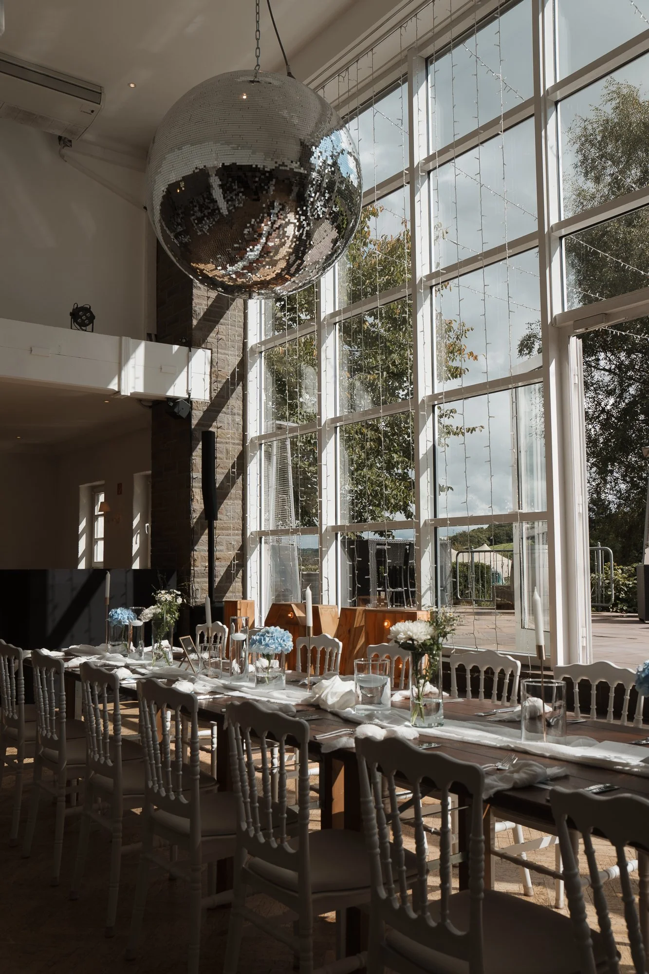 Ein festlich dekorierter Raum mit einem großen Discokugel, langen gedeckten Tischen mit Kerzen, Blumen und Geschirr, vor großen Fenster mit Sonnenlicht und Blick nach draußen.