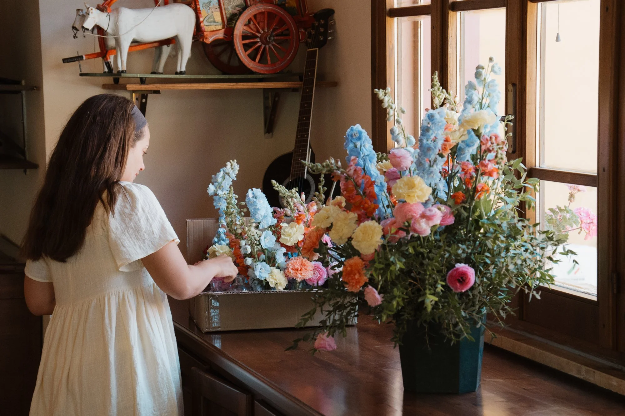 Ein junges Mädchen in einem weißen Kleid stellt einen großen bunten Blumenstrauß auf einer Holzkommode bei einem Fenster mit offenen Holzläden zusammen.