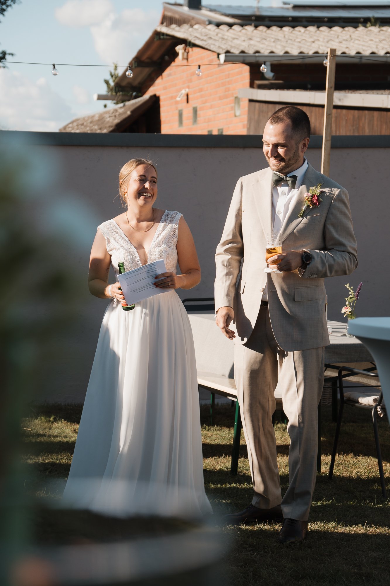 Ein Brautpaar bei einer Hochzeit im Garten, die Braut hält eine Rede und lächelt, der Bräutigam trägt einen grauen Anzug, hält ein Glas und hört zu.