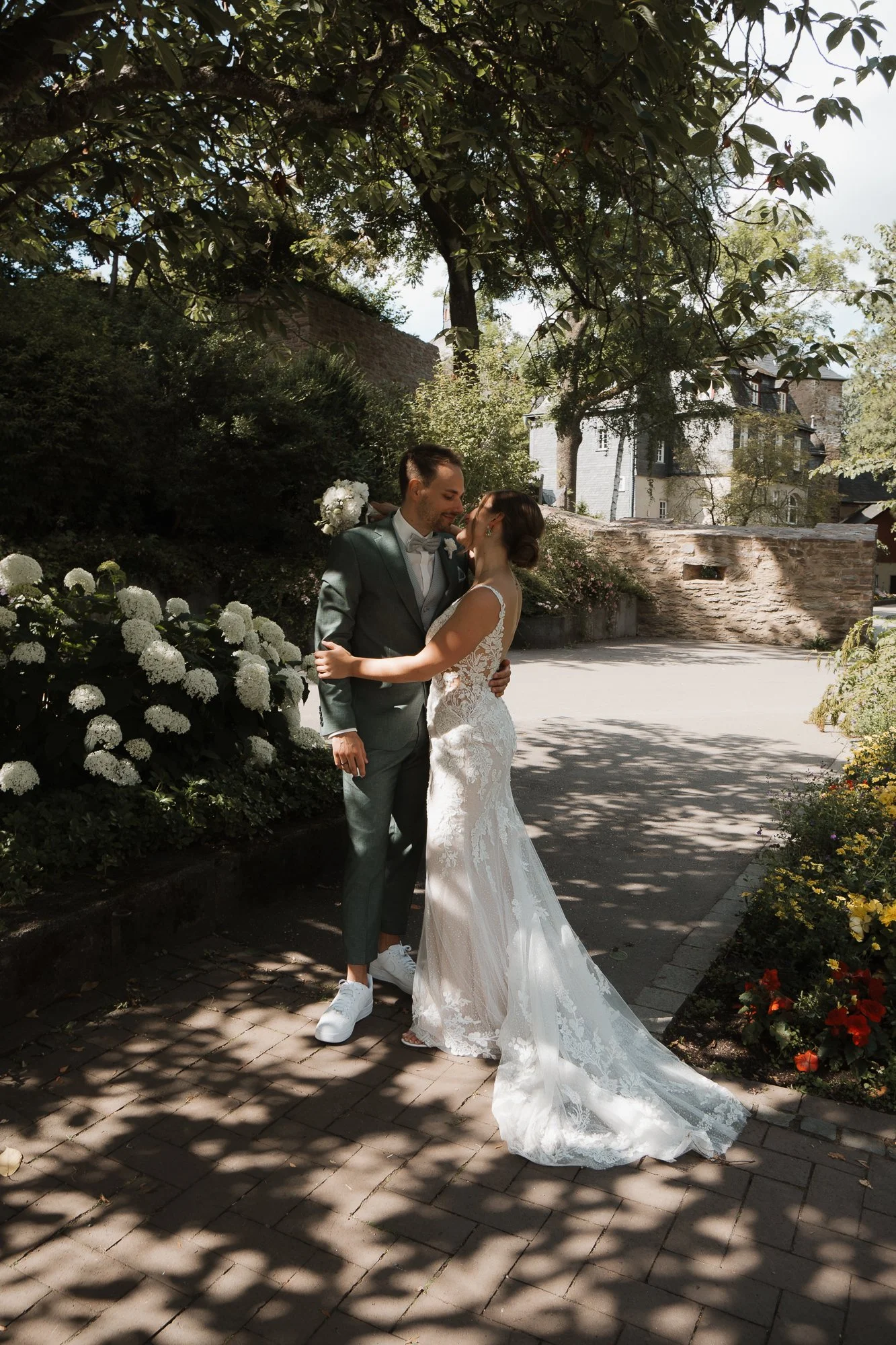 Ein Brautpaar in Hochzeit Kleidung teilt einen Kuss im Schatten eines Baumes in einem Garten mit weißen und gelben Blumen.