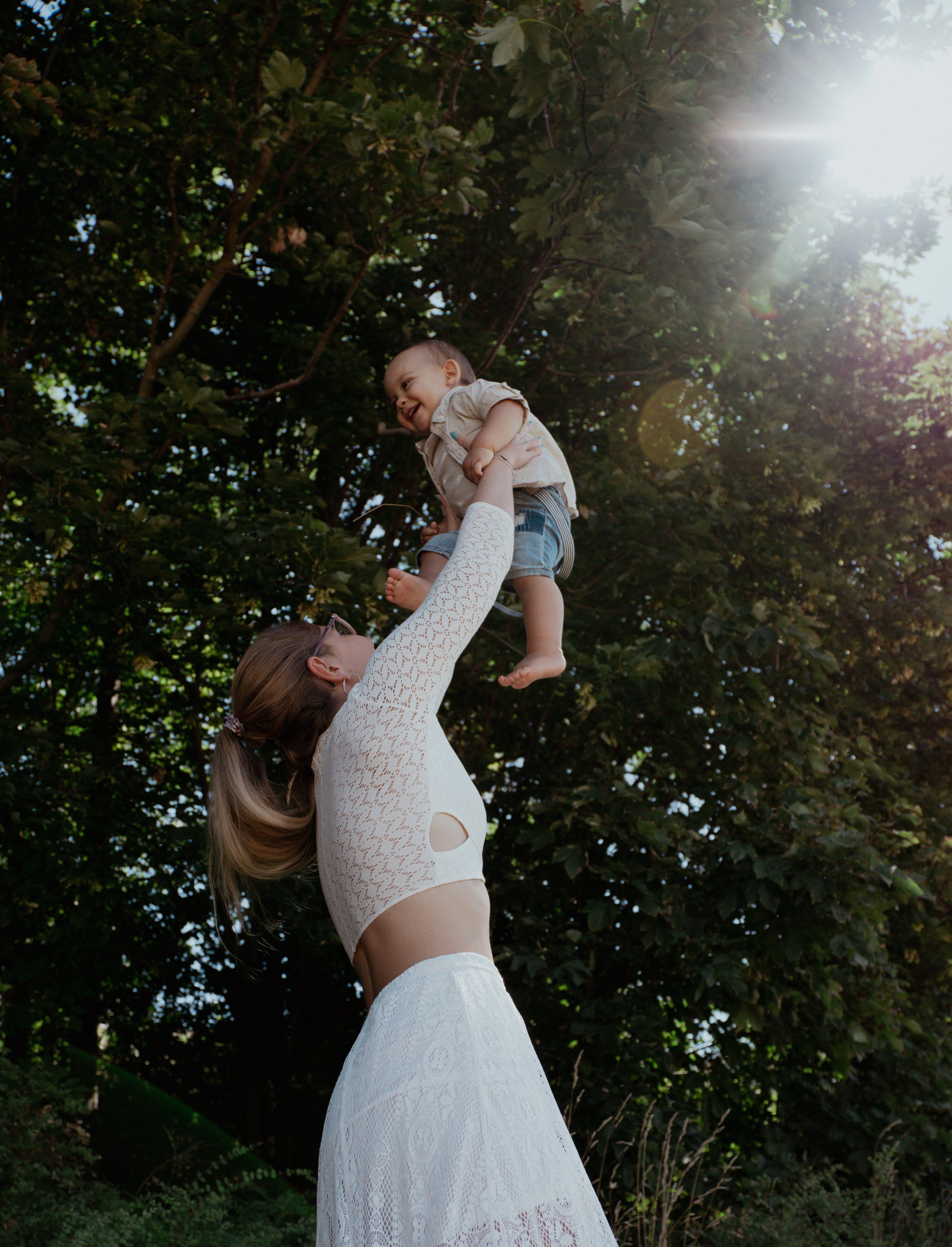 Frau hebt lachendes Kleinkind in den Himmel vor grünen Bäumen mit Sonnenlicht. Ev. Spaziergang im Park.
