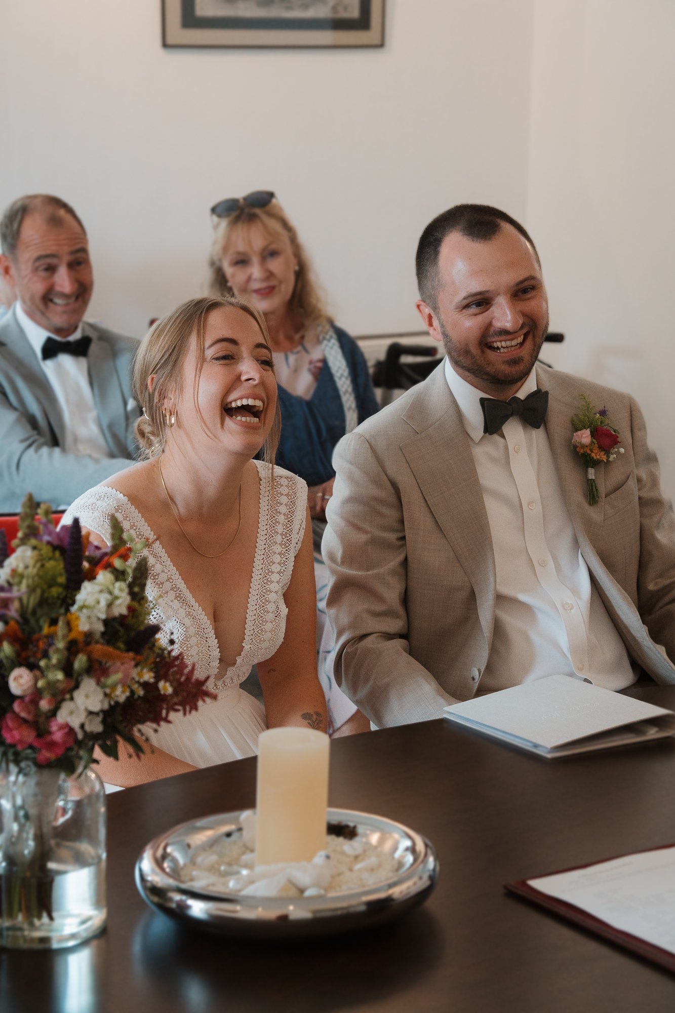 Ein lachendes Brautpaar bei einer Hochzeit, sitzend an einem Tisch mit Blumenstrauß, Kerze und Dokumenten, umgeben von freudigen Gästen.