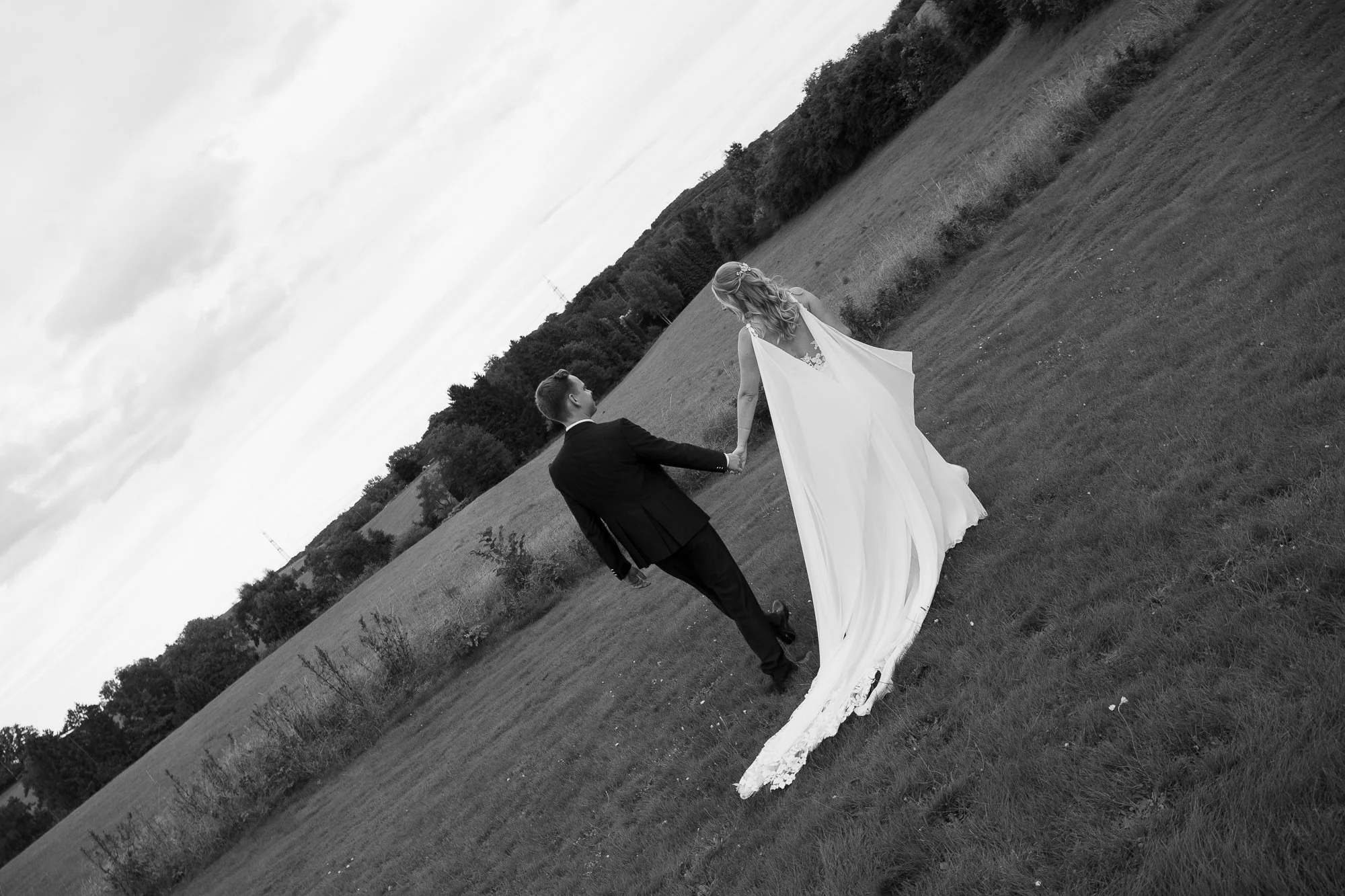 Ein Brautpaar hält Händchen in einem offenen, grünen Feld mit Bäumen im Hintergrund, schwarz-weiß Fotografie.