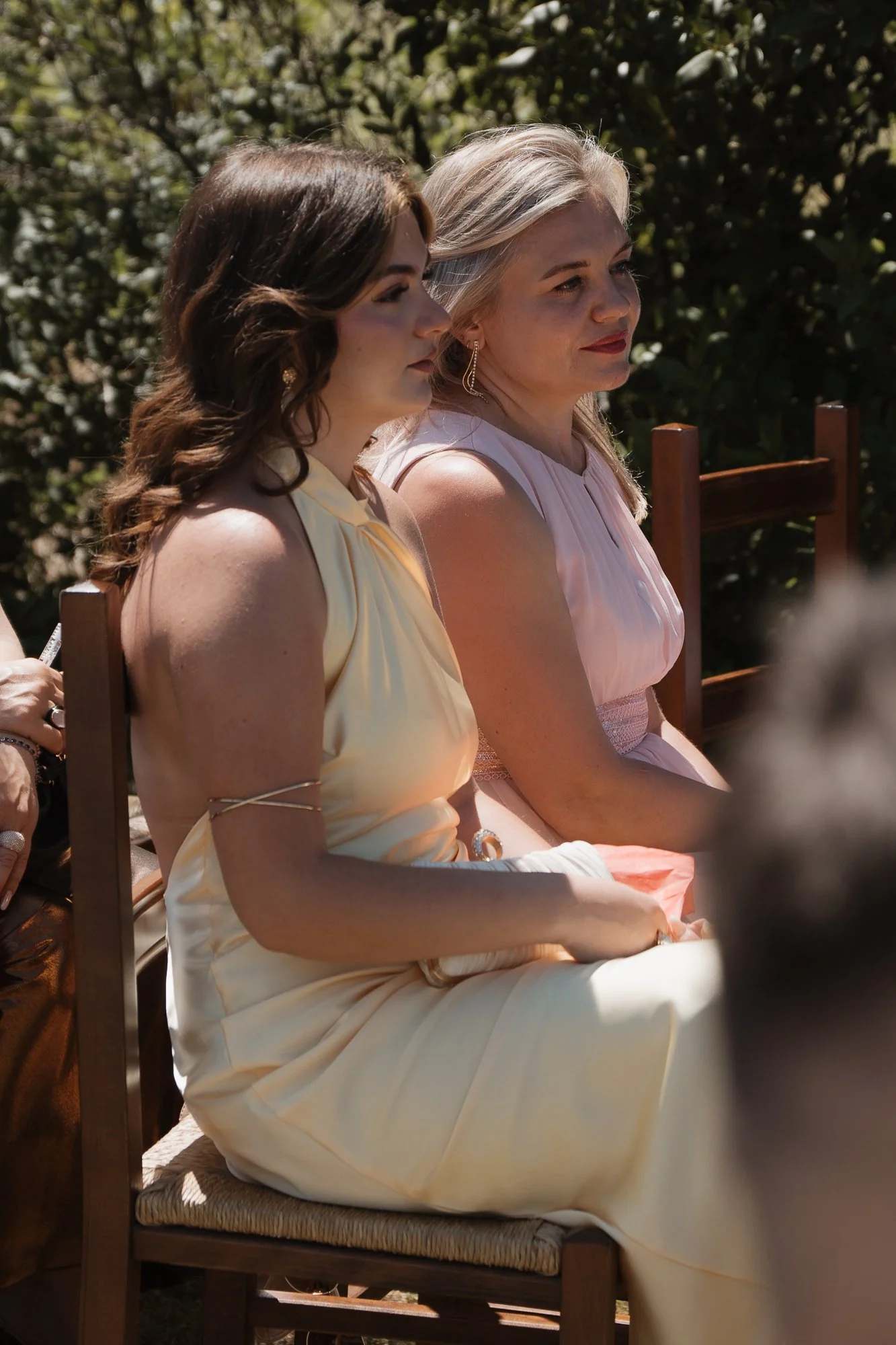 Zwei Frauen sitzen nebeneinander auf Stühlen im Freien, eine trägt ein gelbes Kleid, die andere ein rosa Kleid, und sie schauen aufmerksam in die gleiche Richtung, umgeben von grünen Büschen.