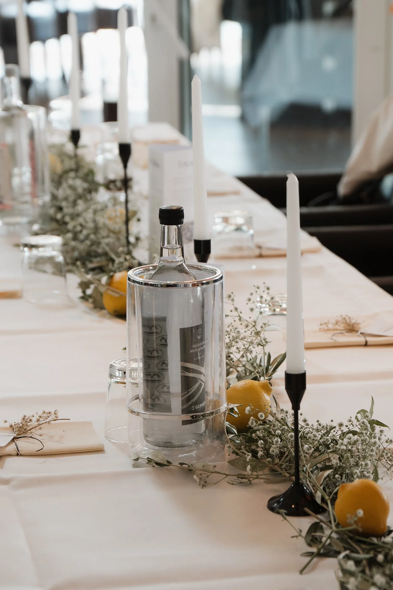 Ein festlich gedeckter Tisch mit weißen Tischtüchern, Kerzen, Zitronen, Blumen und Wasserflaschen, bereit für eine Feier oder Veranstaltung.