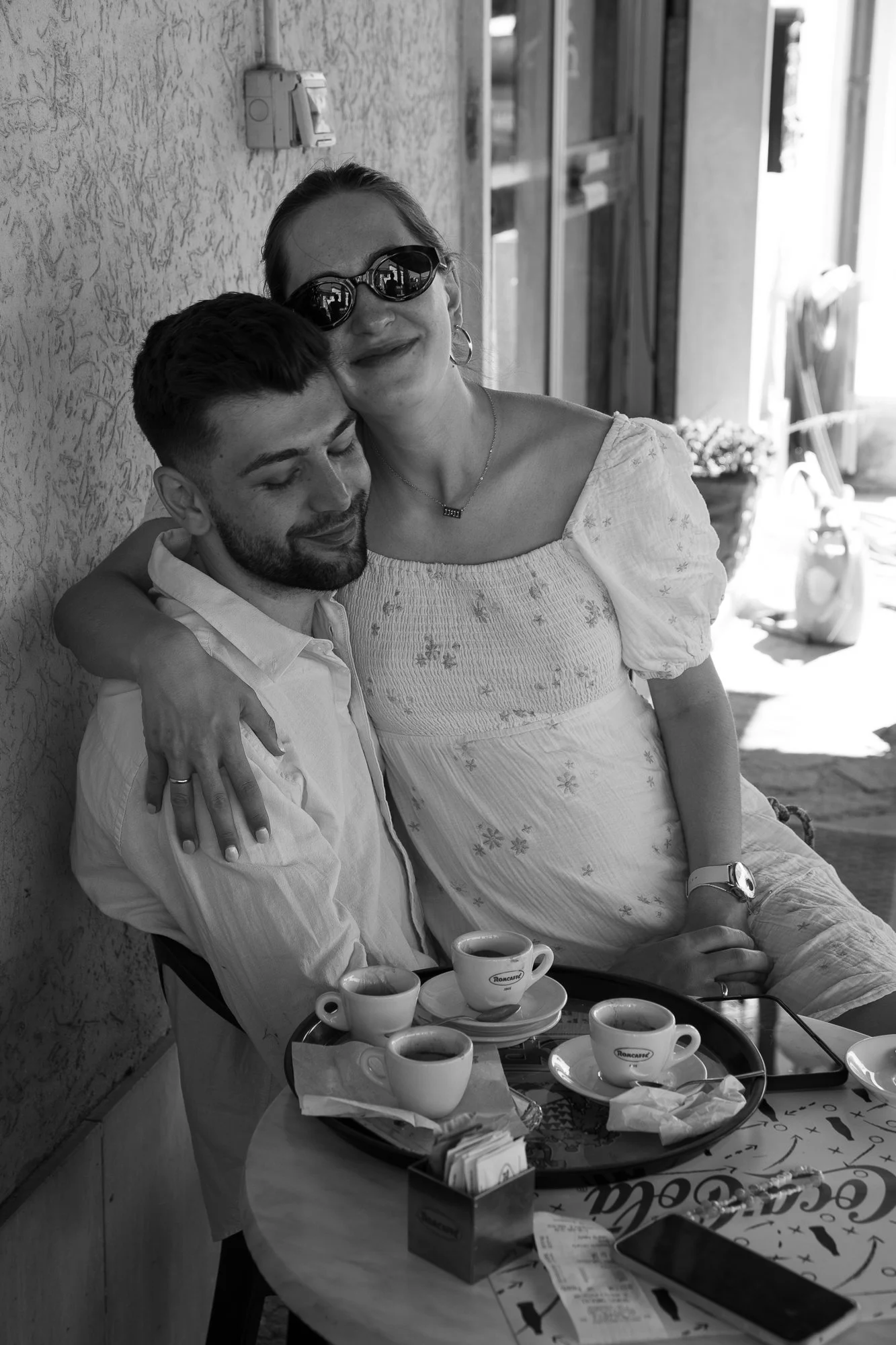 Ein Mann und eine Frau sitzen an einem Tisch, die Frau umarmt den Mann und beide lachen. Es sind Espressotassen auf dem Tisch, und es sieht nach einem Café aus.