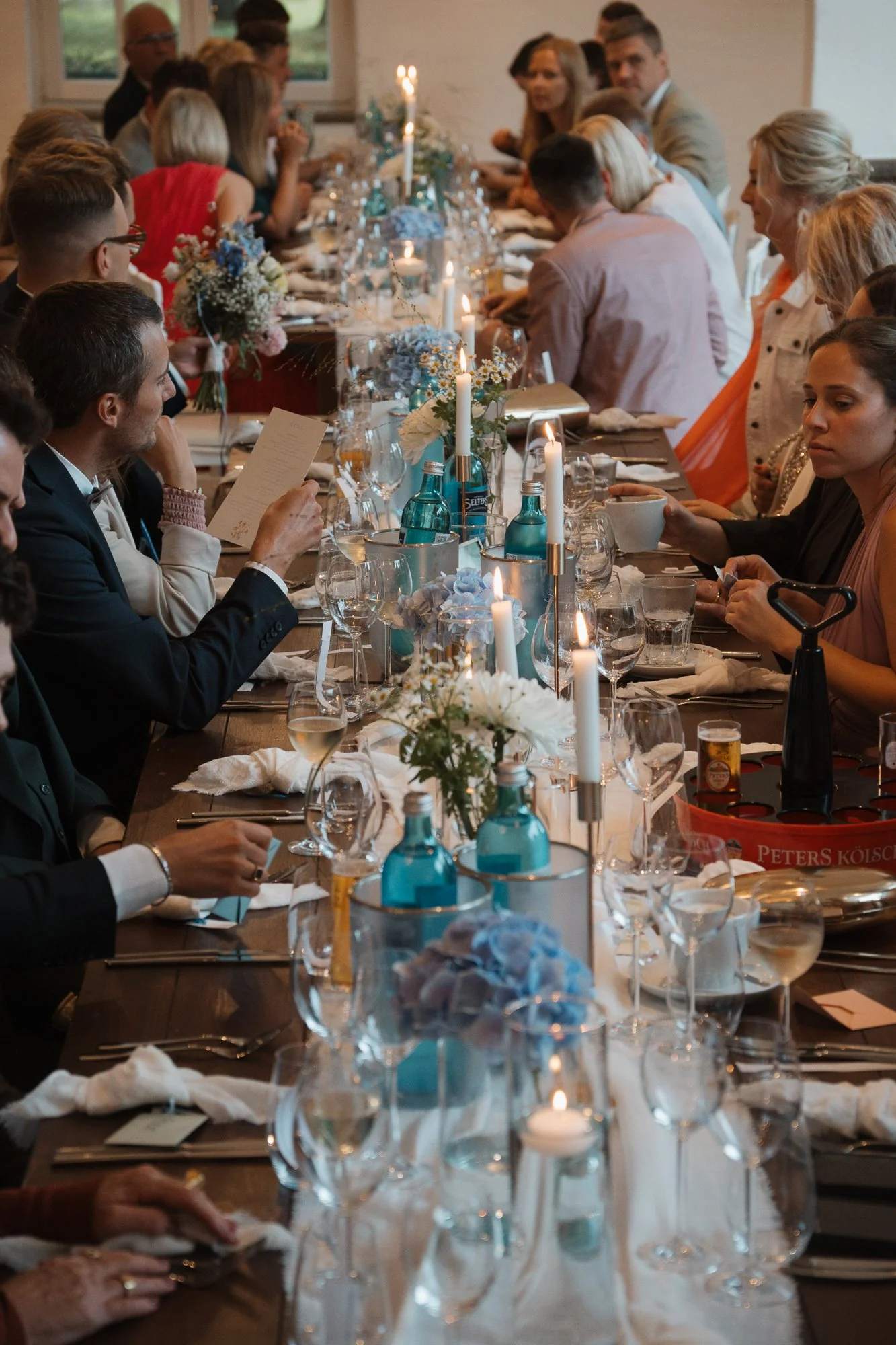 Ein festlich gedeckter langer Esstisch mit Kerzen, Blumen und Getränken, umgeben von Gästen bei einem besonderen Anlass in einem hellen Raum.