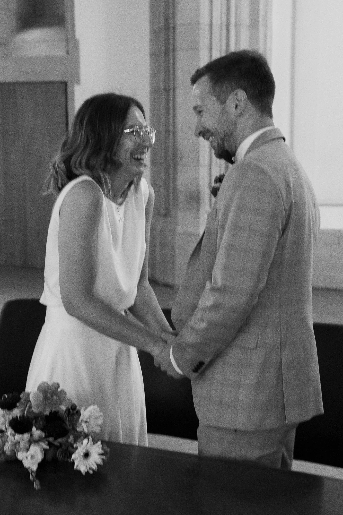 Ein Paar bei einer Hochzeit hält Händchen und grinst sich an, im Hintergrund ein Blumenstrauß auf einem Tisch.