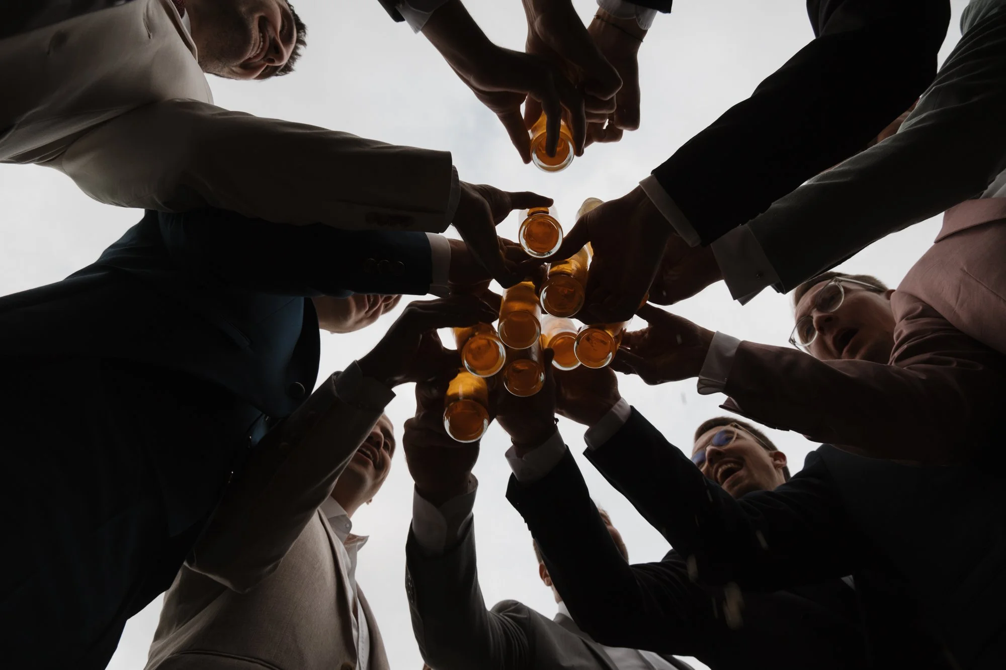 Menschen in Anzügen stoßen mit Biergläsern in einem Kreis zusammen, fotografiert aus der Perspektive nach oben gegen den Himmel.