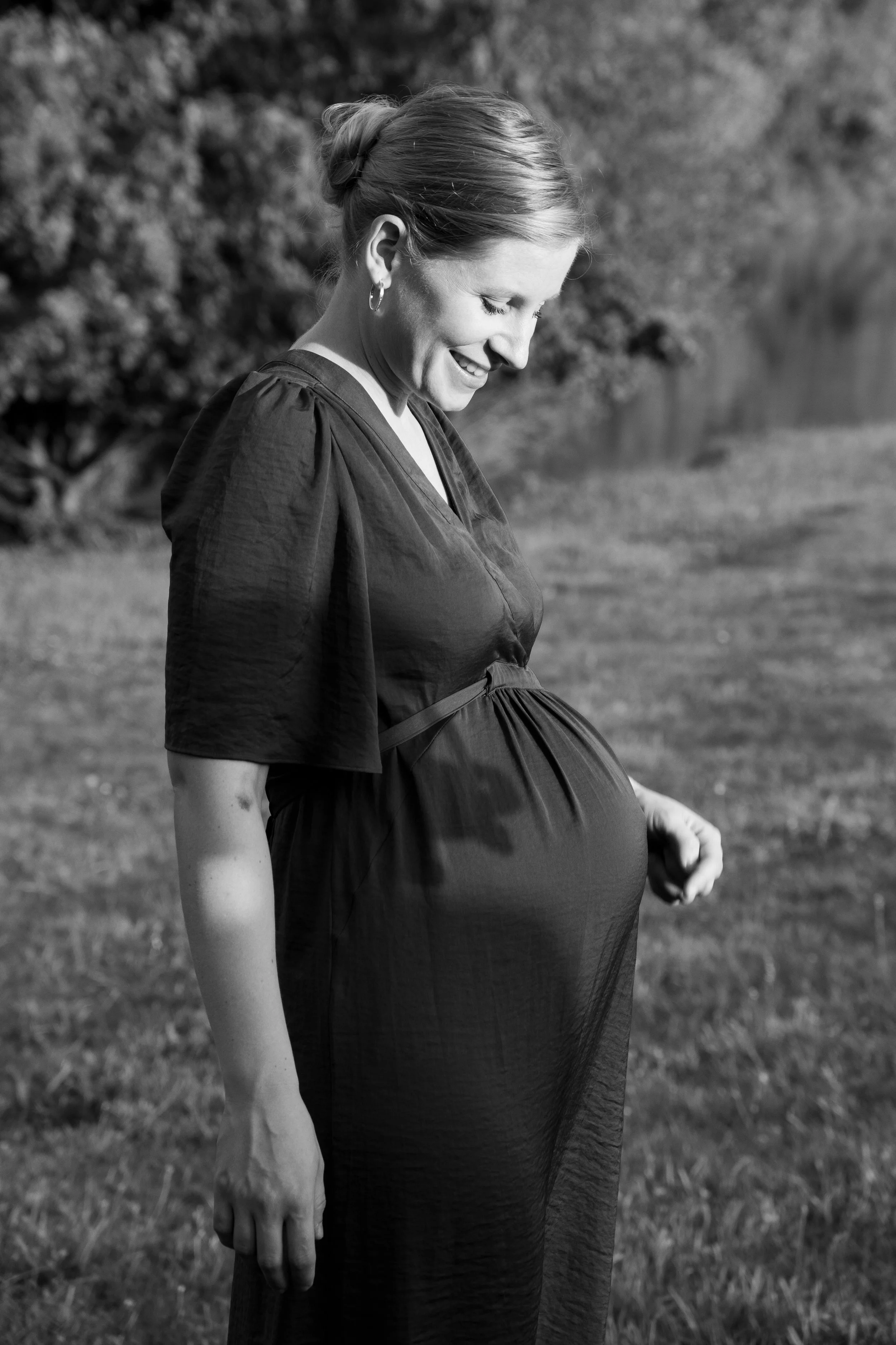 Schwangere Frau in einem schwarzen Kleid, die in einem Park steht und nach unten lächelt.