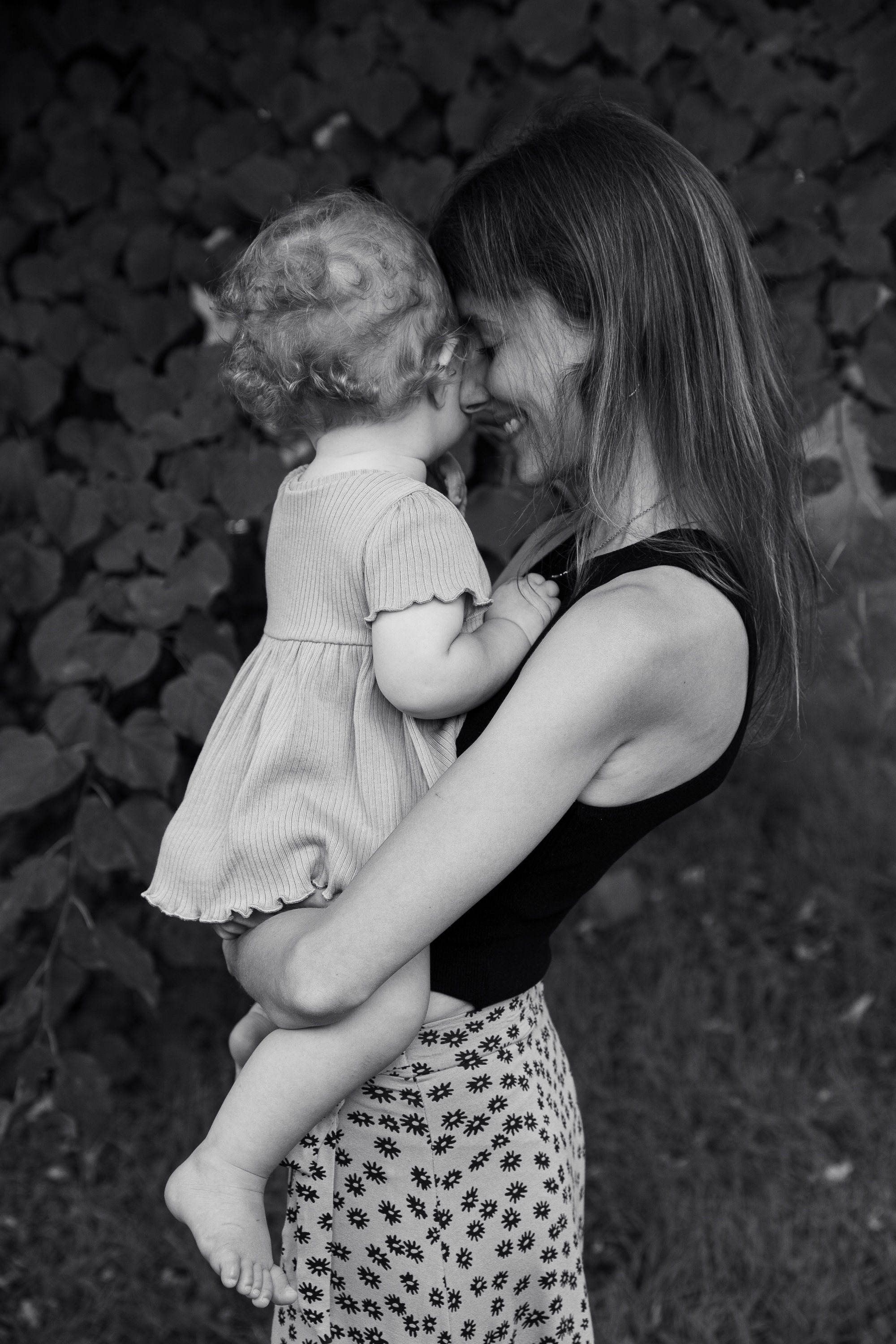 Eine Frau hält ein kleines Kind im Arm, beide lachen und berühren sich mit der Stirn. Im Hintergrund Büschen und Gras, Schwarz-Weiß-Fotografie.