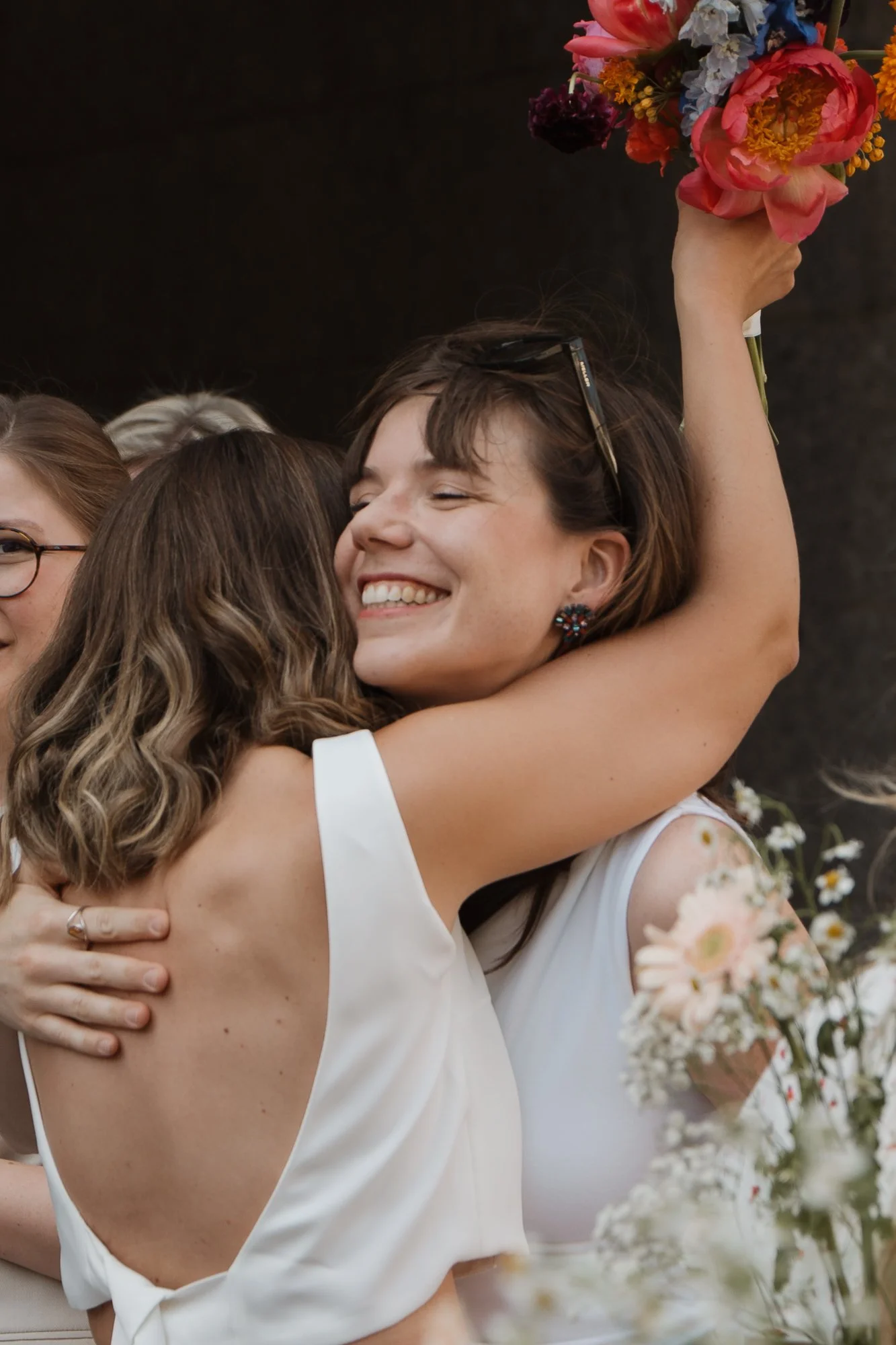 Zwei Frauen umarmen sich bei einer Feier, eine hält einen bunten Blumenstrauß hoch, beide lächeln glücklich.