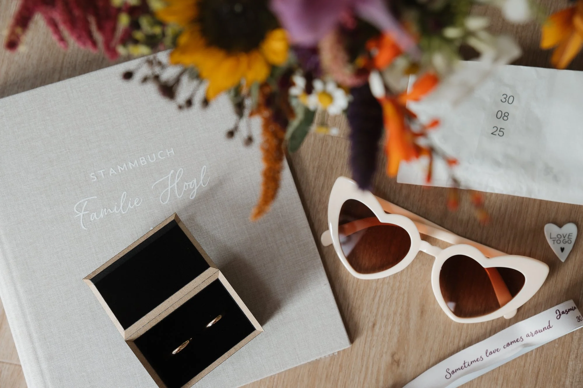 Ein Album mit dem Titel 'Stammbuch Familie Högel' liegt auf einem Holztisch, daneben ein Paar weiße Herz-Sonnenbrillen, ein kleines Geschenkbox mit Ringen, eine Blumensträuße und eine kleine Nachricht mit dem Text 'Sometimes love comes around' sowie 