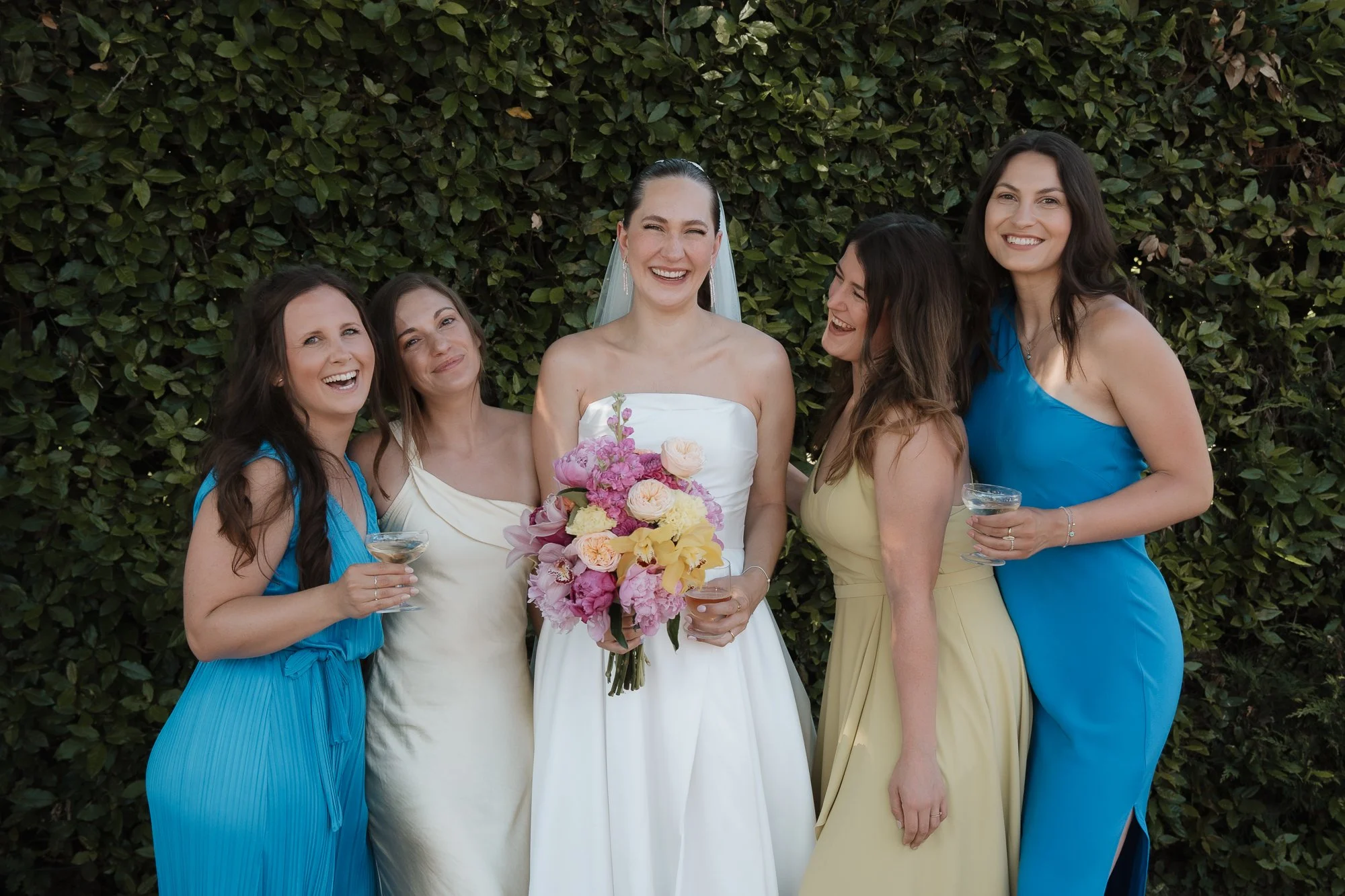 Fünf Frauen in bunten Kleidern lachend bei einer Hochzeit vor einer grünen Hecke, eine hält einen Blumenstrauß, zwei halten Champagner-Gläser.