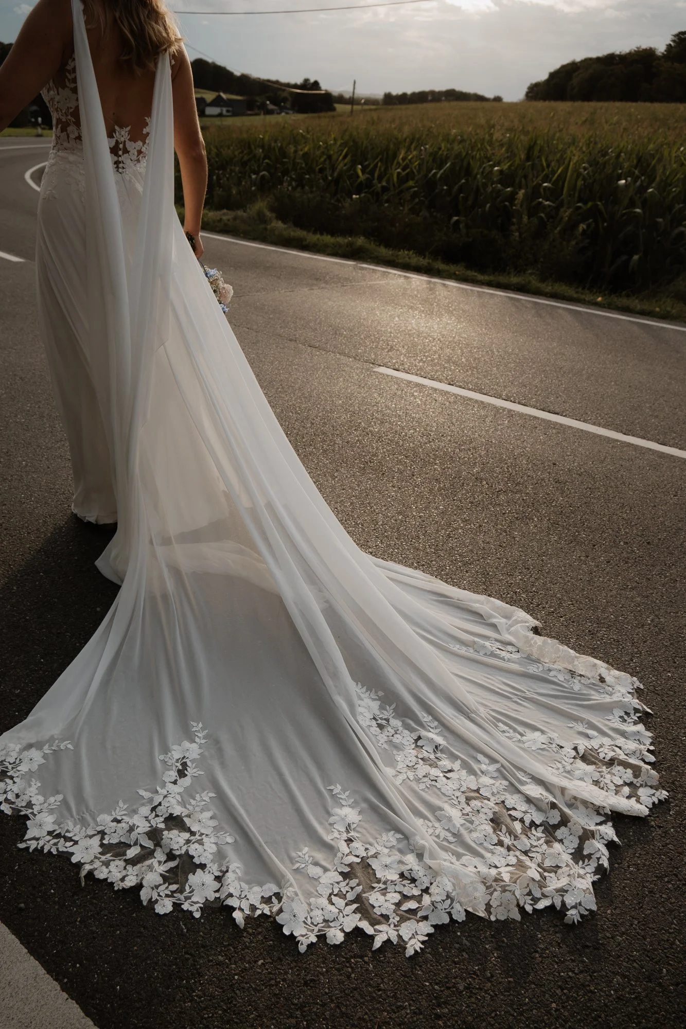 Braut in Hochzeitskleid mit langer Schleppe auf Asphaltstraße in ländlicher Umgebung bei Sonnenuntergang.