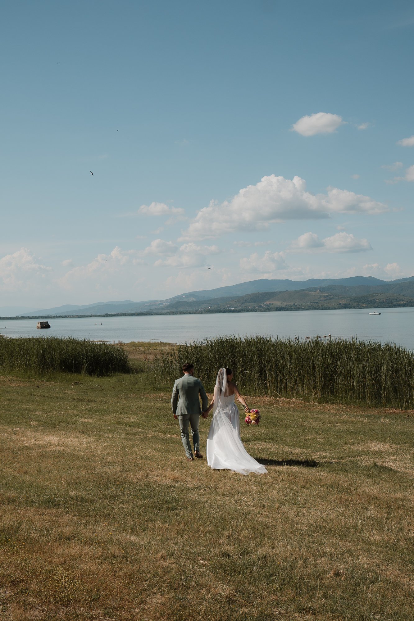 Ein Brautpaar läuft Hand in Hand an einem Seeufer bei schönem Wetter, die Braut trägt ein weißes Kleid und hält einen bunten Blumenstrauß, im Hintergrund sind Berge und Wolken am Himmel zu sehen.