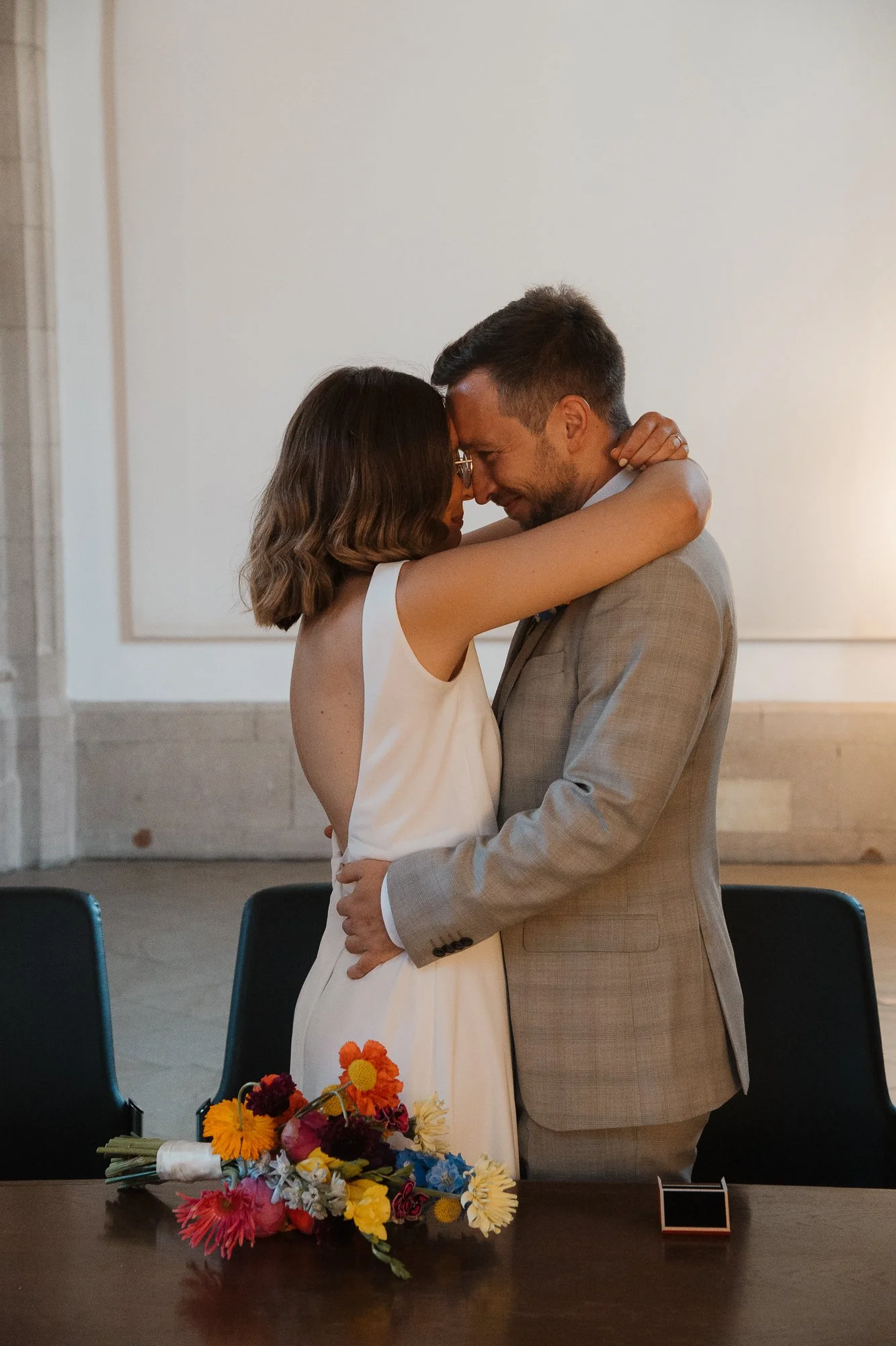 Ein Paar umarmt sich in einer modernen, eleganten Umgebung, wahrscheinlich bei einer Heiratszeremonie oder einem besonderen Anlass, mit einem bunten Blumenstrauß auf dem Tisch vor ihnen.