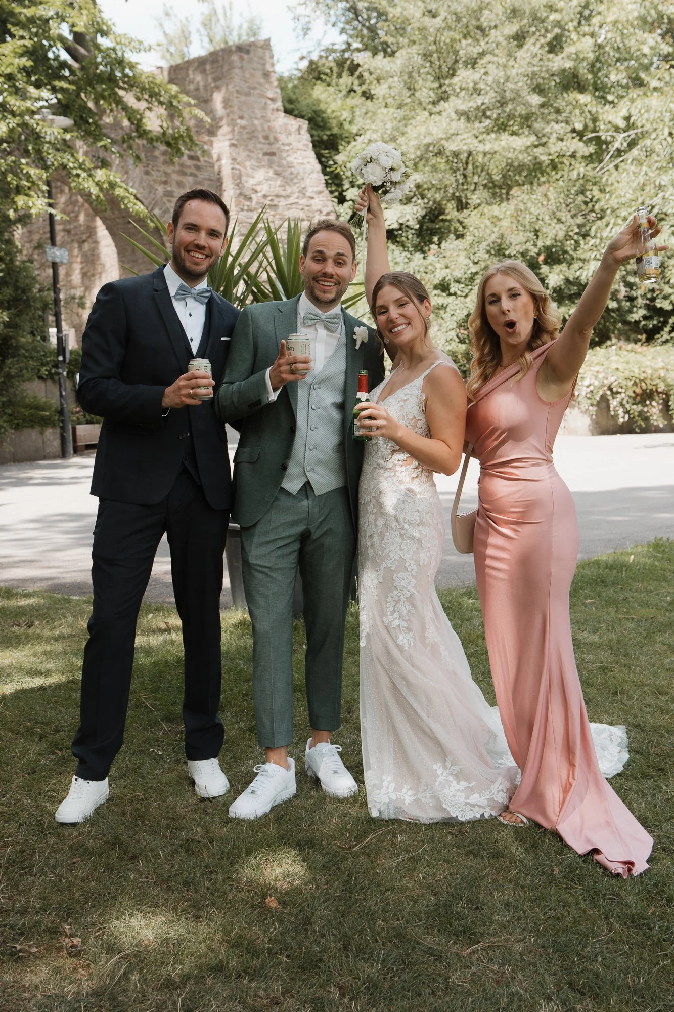 Eine Gruppe von Menschen, darunter ein Brautpaar, die bei einer Hochzeit im Freien fröhlich feiern, mit einigen Getränken in der Hand, umgeben von Bäumen und einer alten Steinmauer.