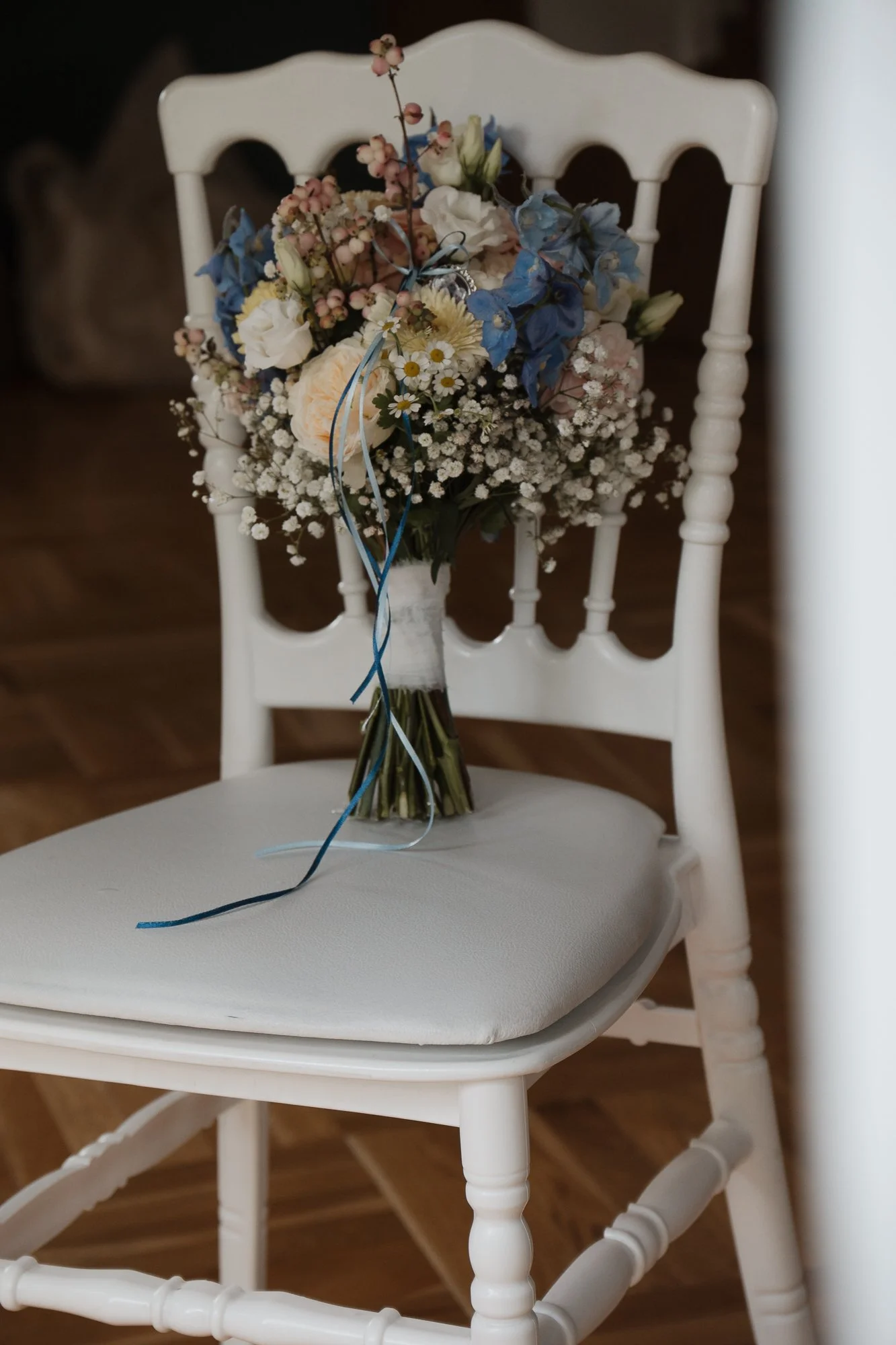 Blumenstrauß auf einem weißen Stuhl, bestehend aus weißen, blauen und rosa Blumen, mit blauer Tannenrinde und Bändern, auf einer gepolsterten Sitzfläche.