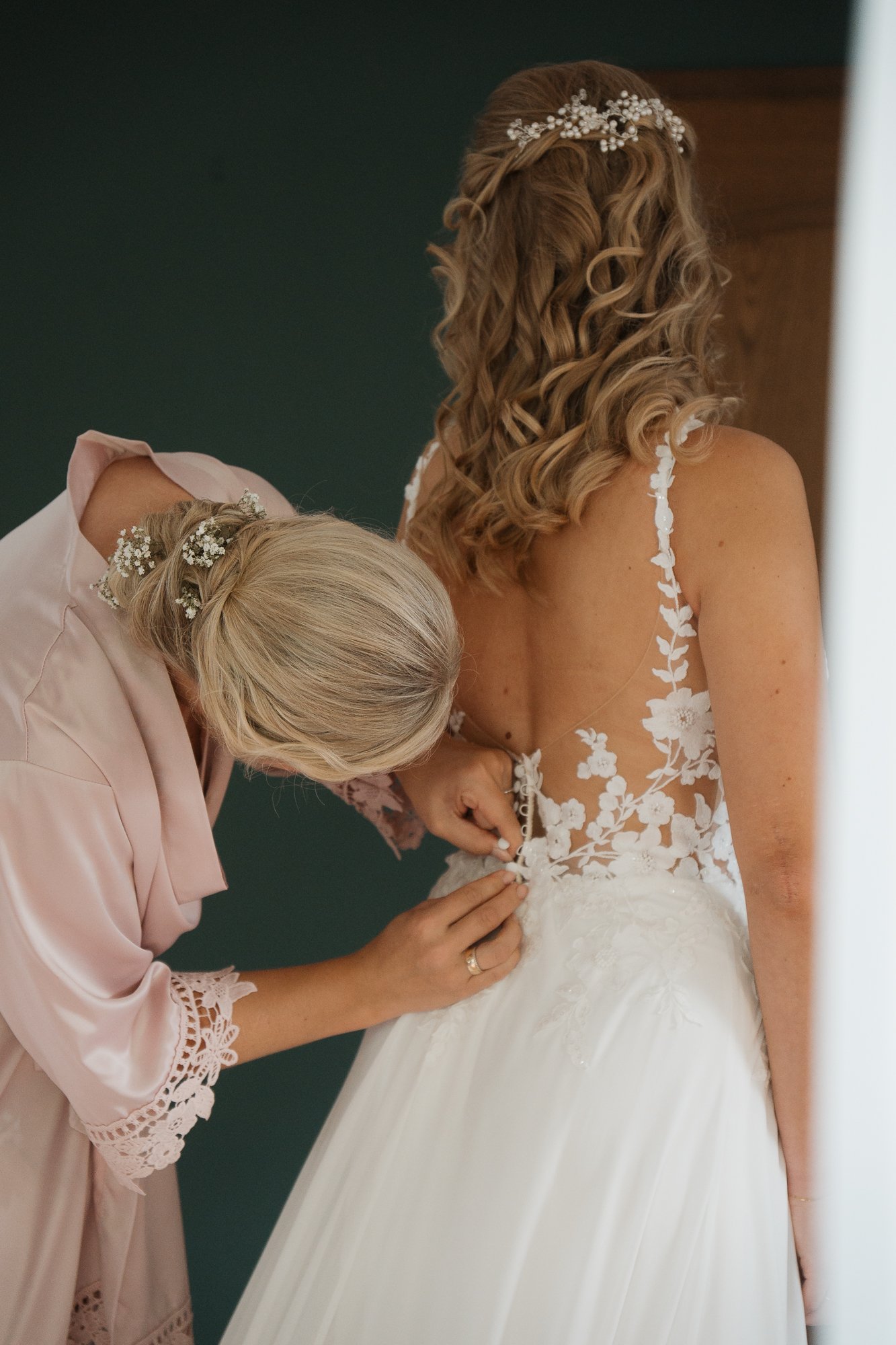 Eine Braut wird geholfen, den Verschluss ihres weißen Hochzeitskleides zu schließen, während eine Frau sie dabei unterstützt.