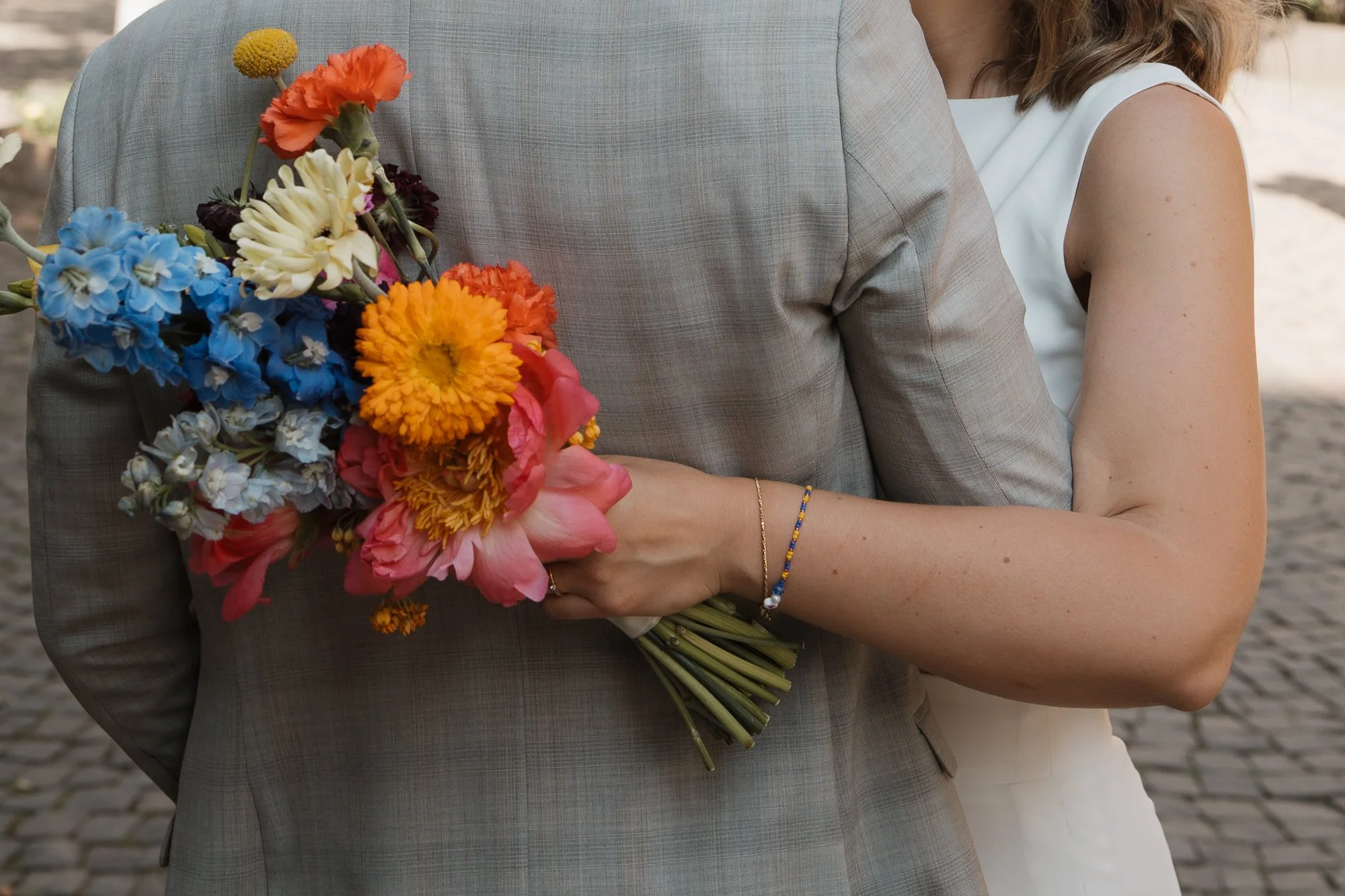 Nahaufnahme eines Paares bei einer Hochzeit, Frau hält einen bunten Blumenstrauß hinter dem Rücken des Mannes, im Hintergrund eine gepflasterte Straße.